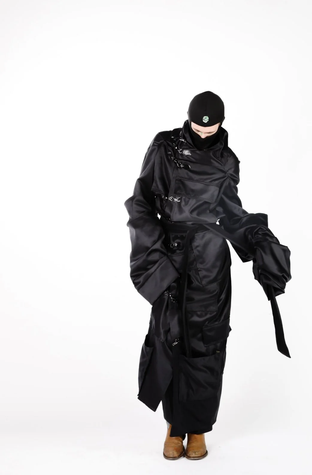 Person dressed in black clothing including a beanie, face covering, jacket, and pants, standing against a plain white background.