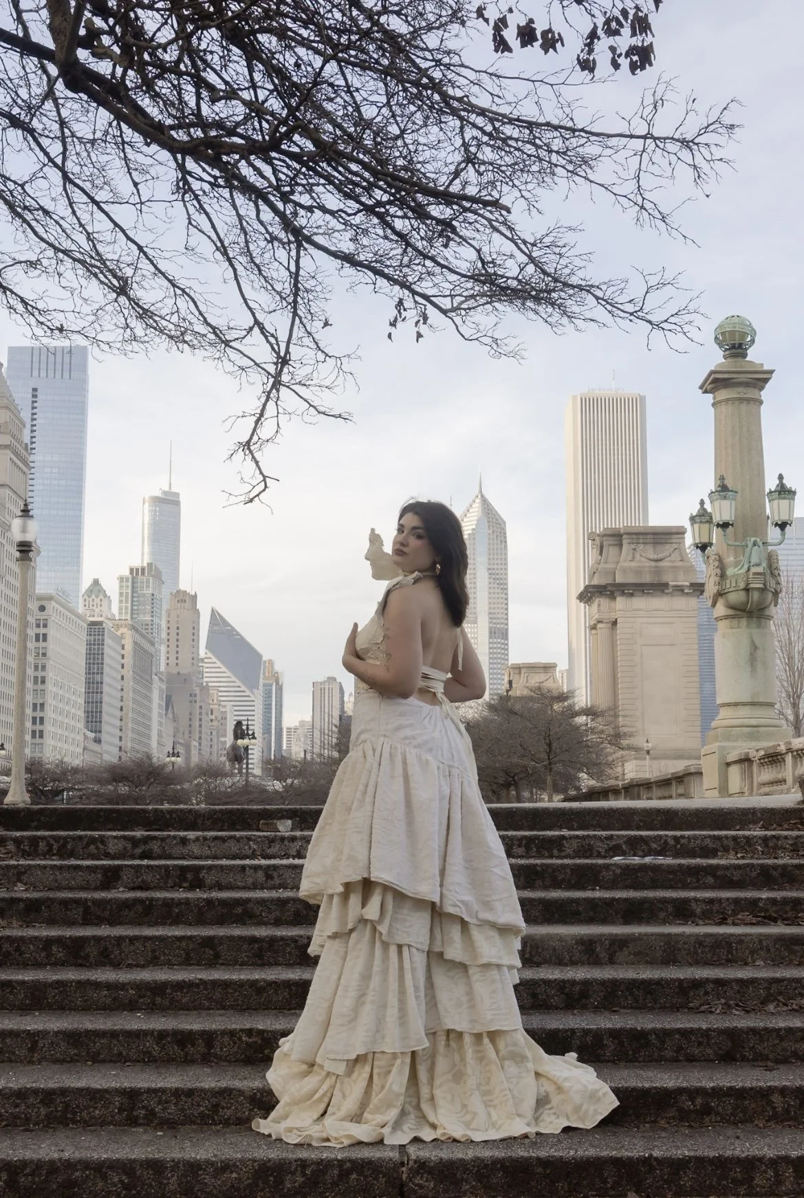 A woman in an elegant, cream-colored Victorian-style gown stands on stone steps in an urban park with a city skyline in the background, including tall buildings and leafless trees.