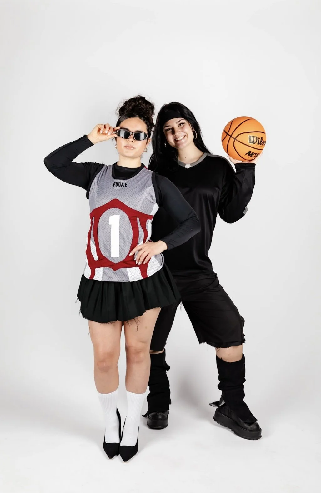 Two women standing against a white background, one wearing a basketball jersey, skirt, and sunglasses, and the other holding a basketball and wearing black clothing and boots.