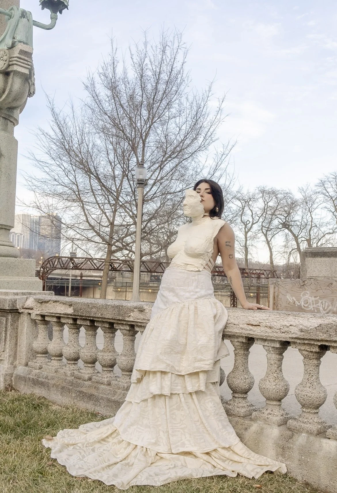 A woman in a vintage-style cream dress with a layered skirt stands outdoors, leaning against a stone railing, with trees and a cityscape in the background. She has dark hair and tattoos on her arm, and is holding a piece of fabric or paper near her f