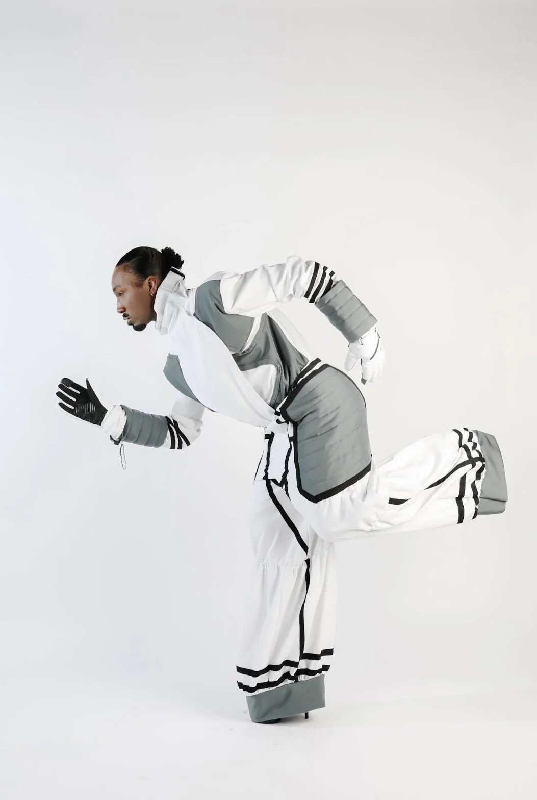 A person dressed in white and gray sportswear, balancing on one leg with the other leg extended behind, running or in a dance pose against a plain white background.