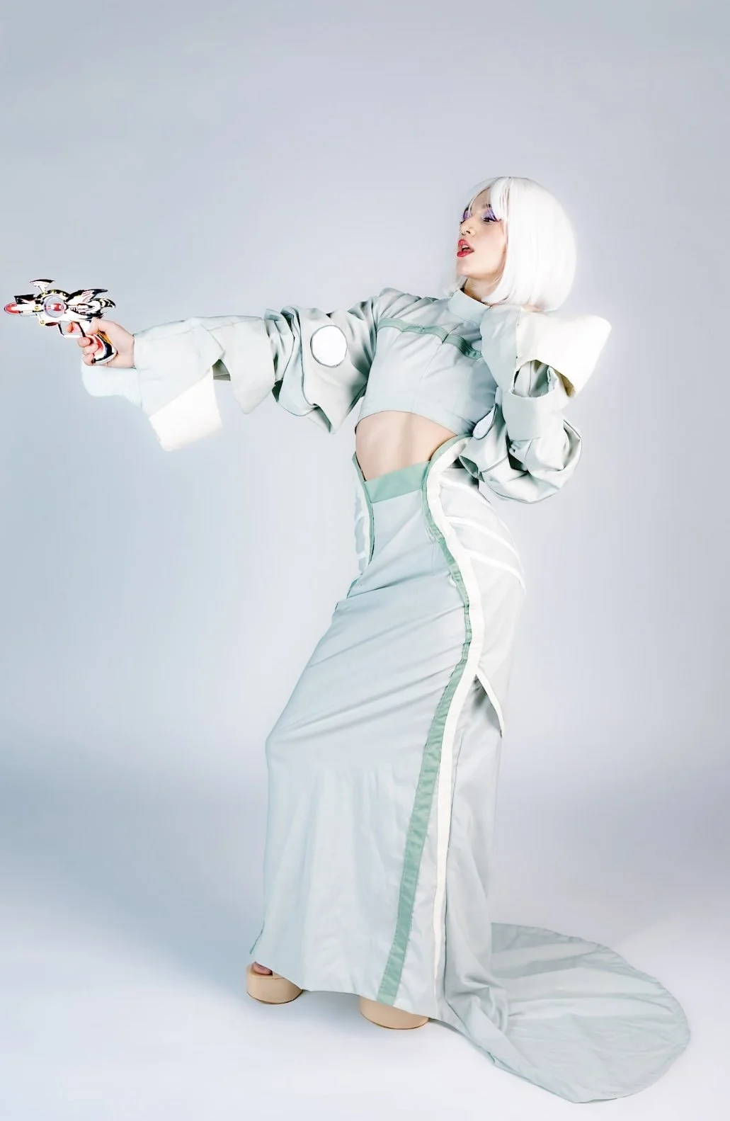 A woman dressed in a futuristic silver outfit with green accents, holding a laser gun, with short white hair and bright makeup, posing in front of a plain light background.