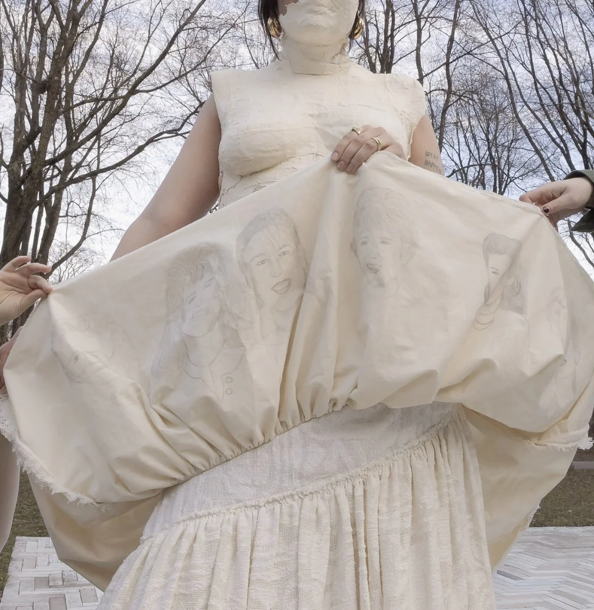Person holding up a cream-colored dress with hand-drawn portraits of women on the fabric, outdoors with leafless trees in the background.