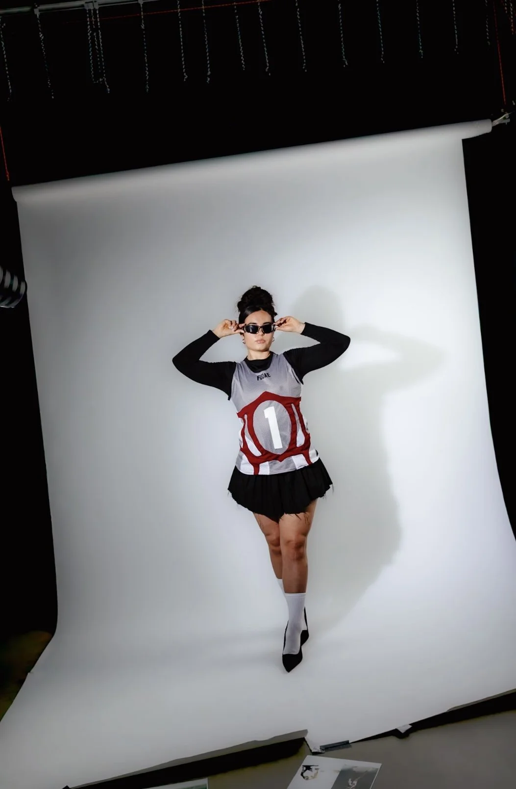 A woman standing in a photo studio, wearing sunglasses, a sporty shirt with the number 1 on it, a black skirt, and high socks, posing against a white backdrop.