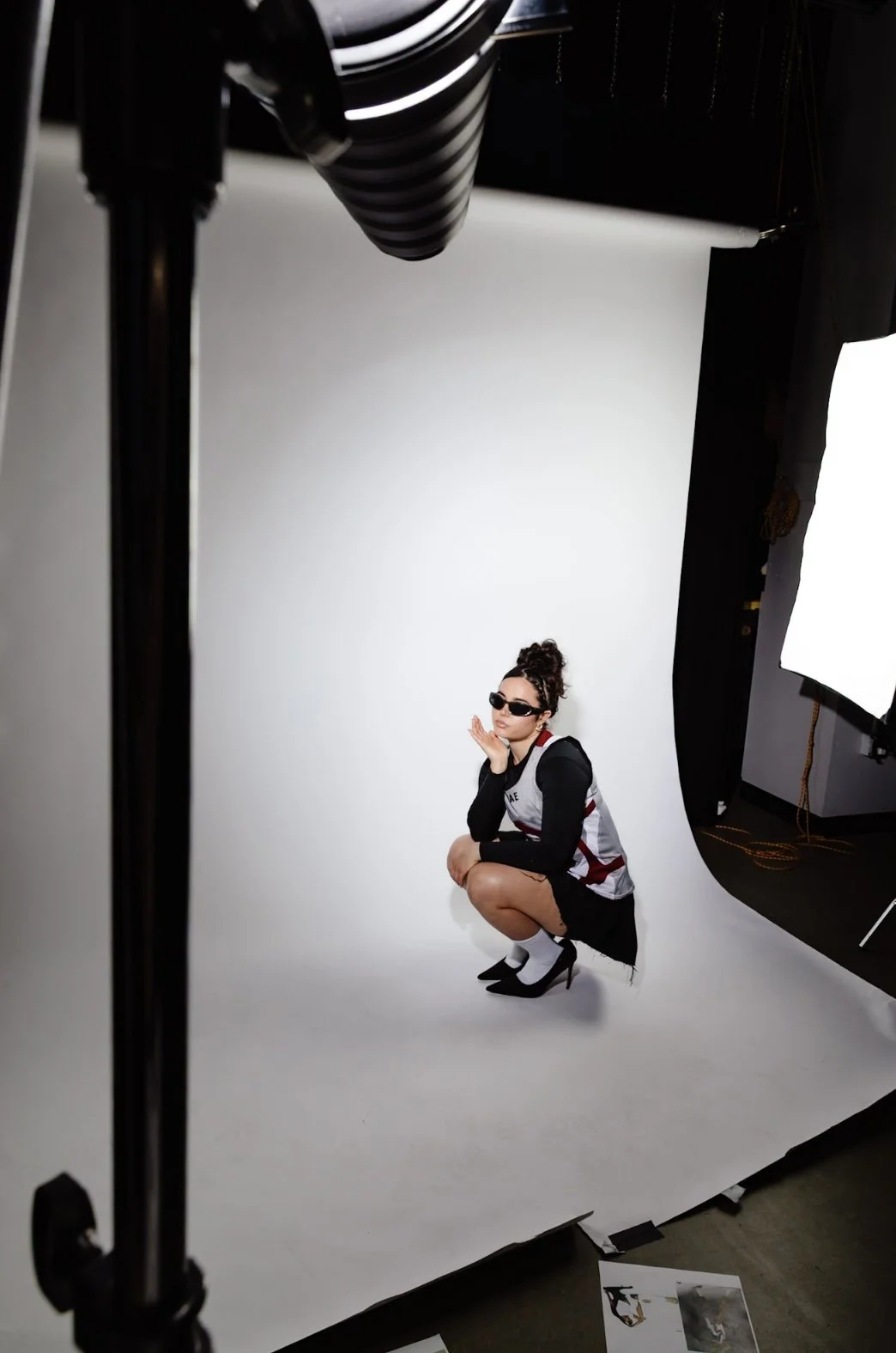 A woman wearing sunglasses, high heels, and a black and white outfit, posing in a squatting position against a white backdrop in a photography studio.