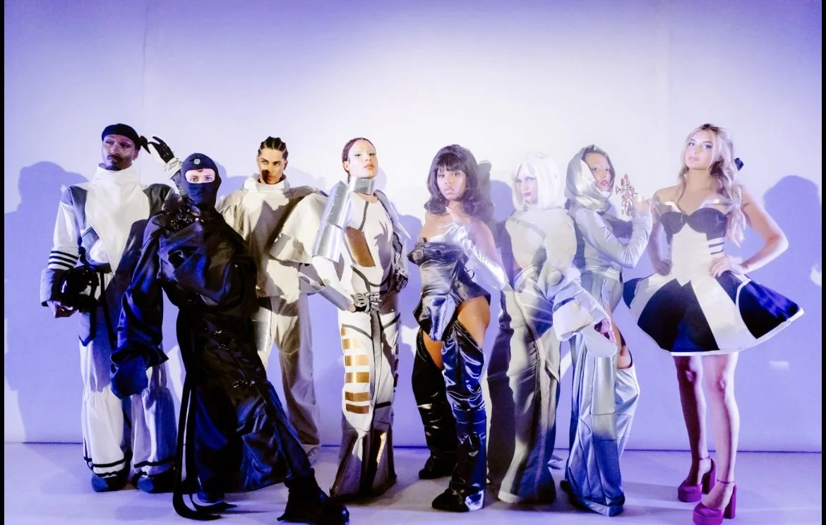 Group of nine models wearing avant-garde fashion with futuristic and artistic designs poses against a plain white background.