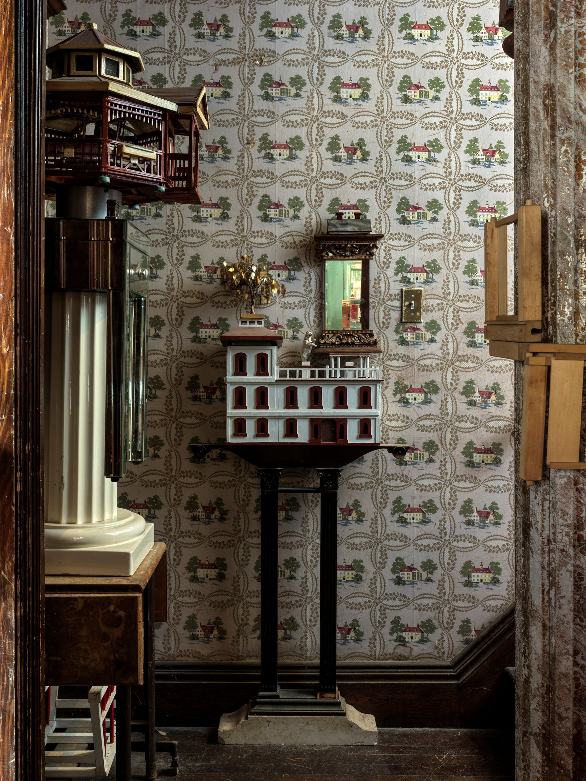 A room with vintage decor featuring patterned wallpaper with houses and trees, a miniature dollhouse on a black pedestal, a small ornate mirror, a miniature white and red building, and various antique furniture and decorations.