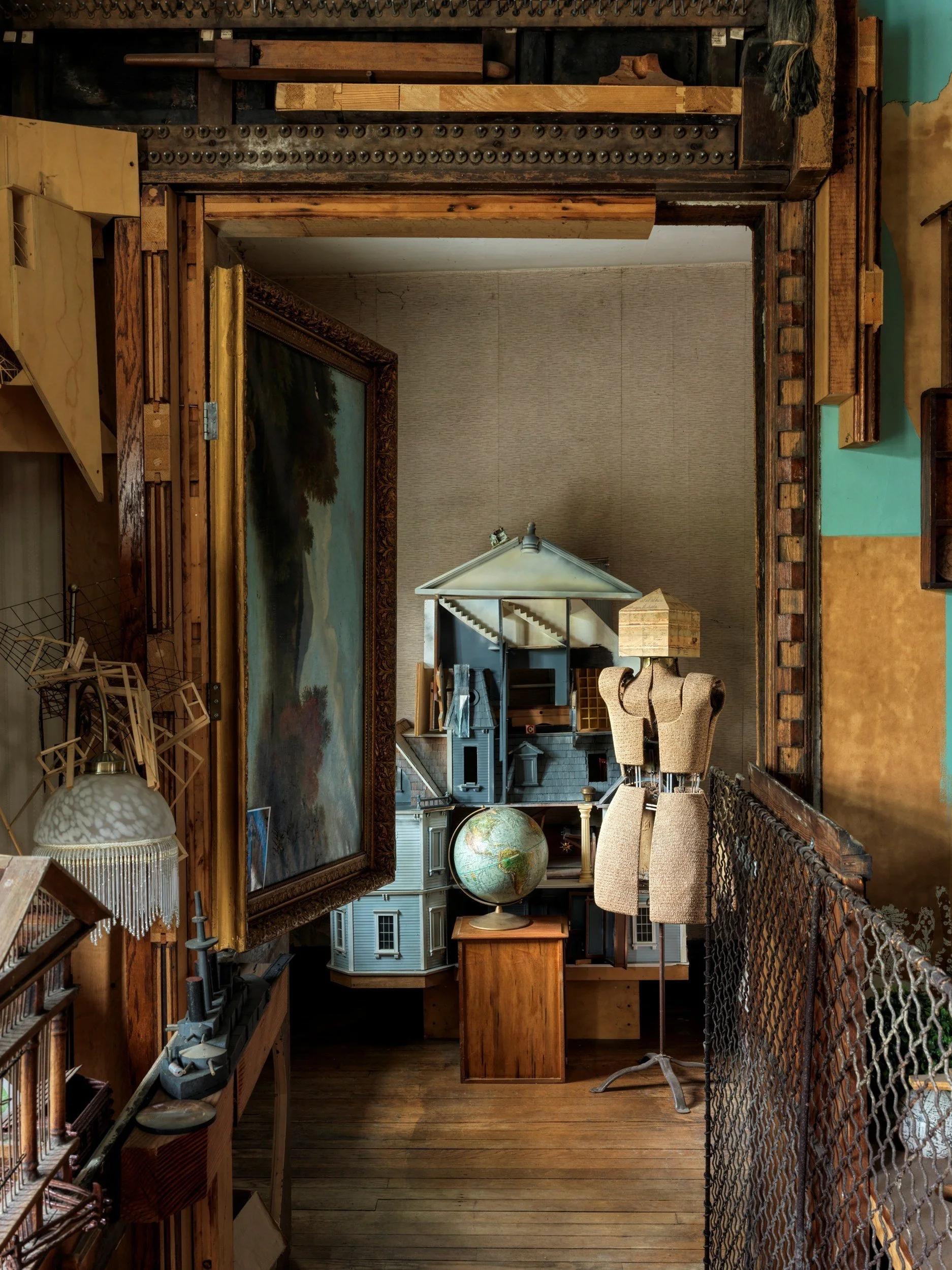 A cluttered room with various furniture and objects, including a large framed landscape painting on the left wall, a globe, a small dollhouse, and a standing lamp, viewed through a wooden and metal railing.