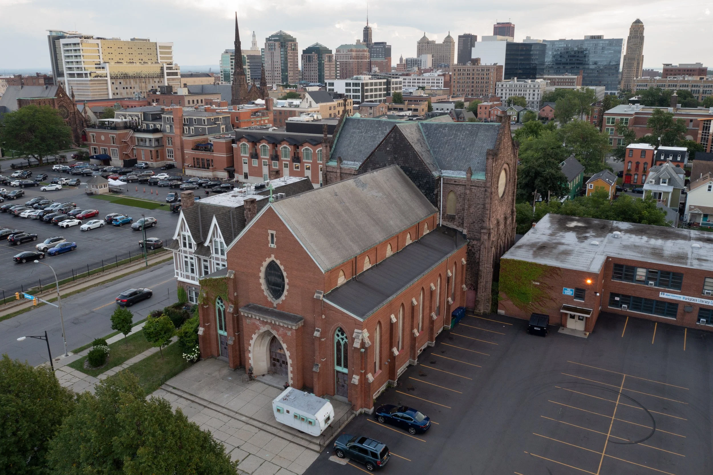 Aerial view of a cityscape with a historic brick church in the foreground, surrounded by parking lots, trees, and a mix of modern and historic buildings in the background under a partly cloudy sky.