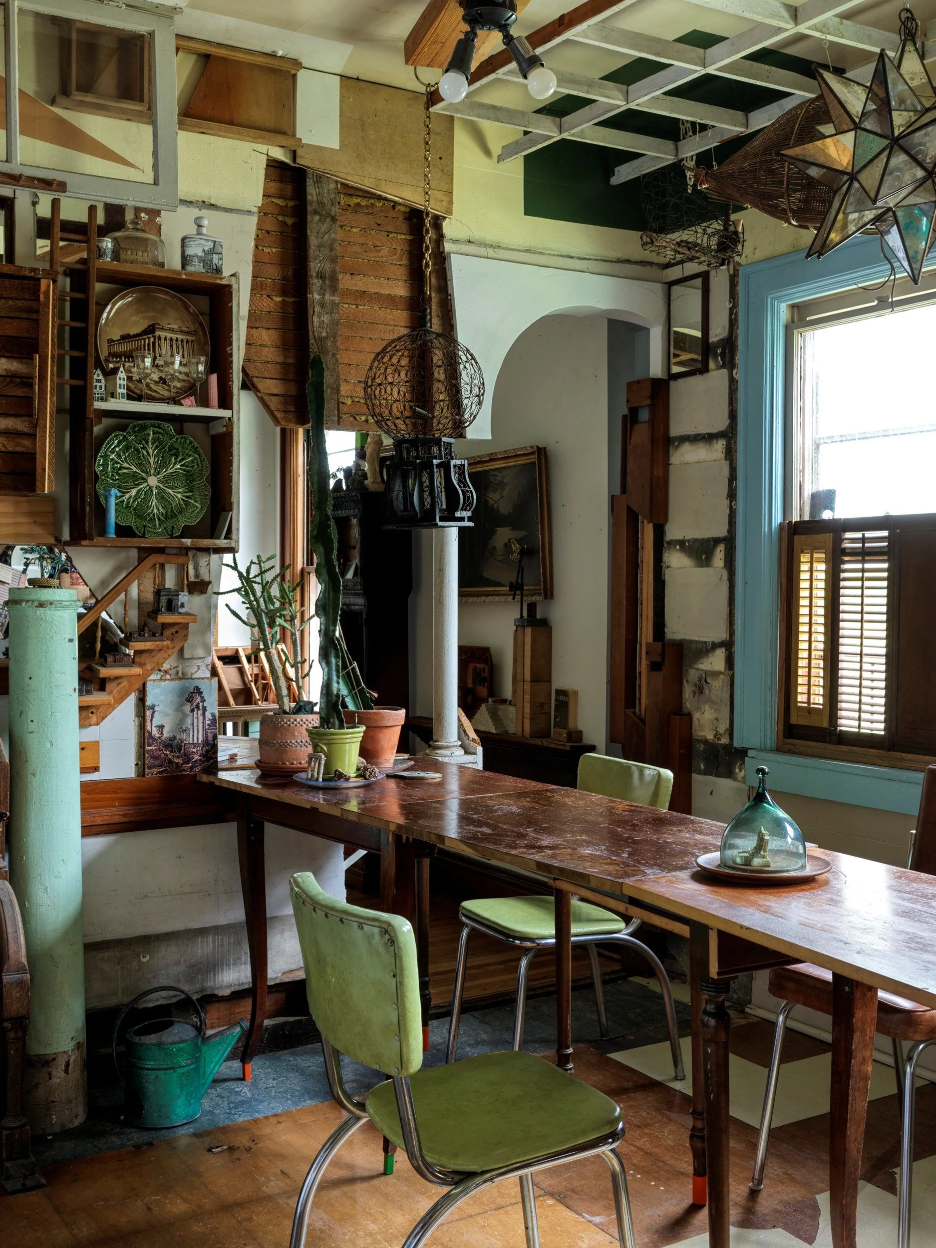 Eclectic interior of a room with a wooden table, vintage green chairs, potted plants, shelves with decorative items, a window with wooden shutters, and hanging lights and decor, featuring rustic and bohemian style elements.