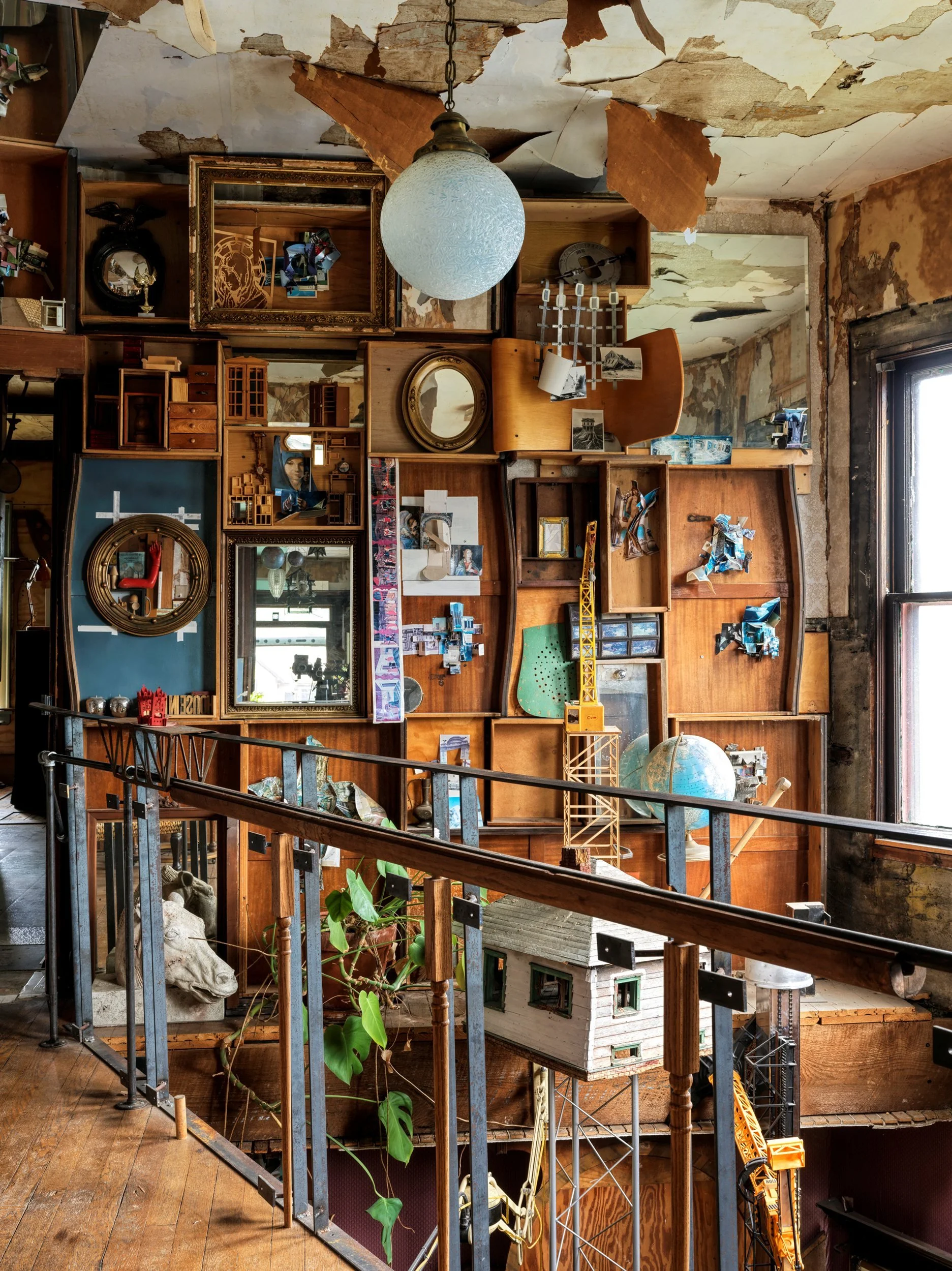 A cluttered indoor space with exposed ceiling paint peeling and various decorative and miniature items on wooden shelves, including a globe, old photographs, a mirror, miniature furniture, construction toy cranes, a plant, a skull sculpture, and vari