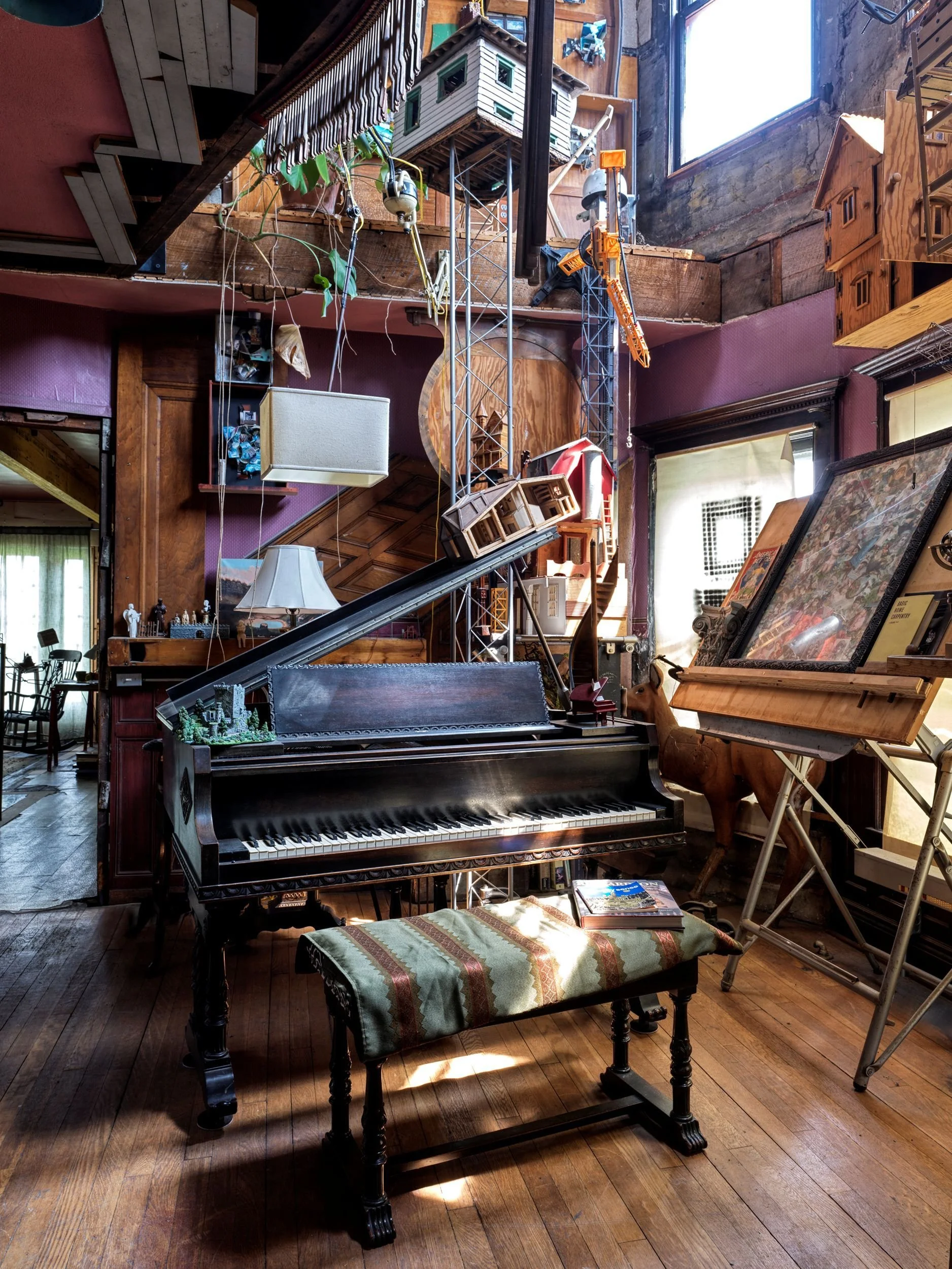 A large grand piano with a floral-patterned upholstered bench in a cozy, well-lit room filled with furniture, model houses, and art supplies, with a skylight and bookshelves visible in the background.