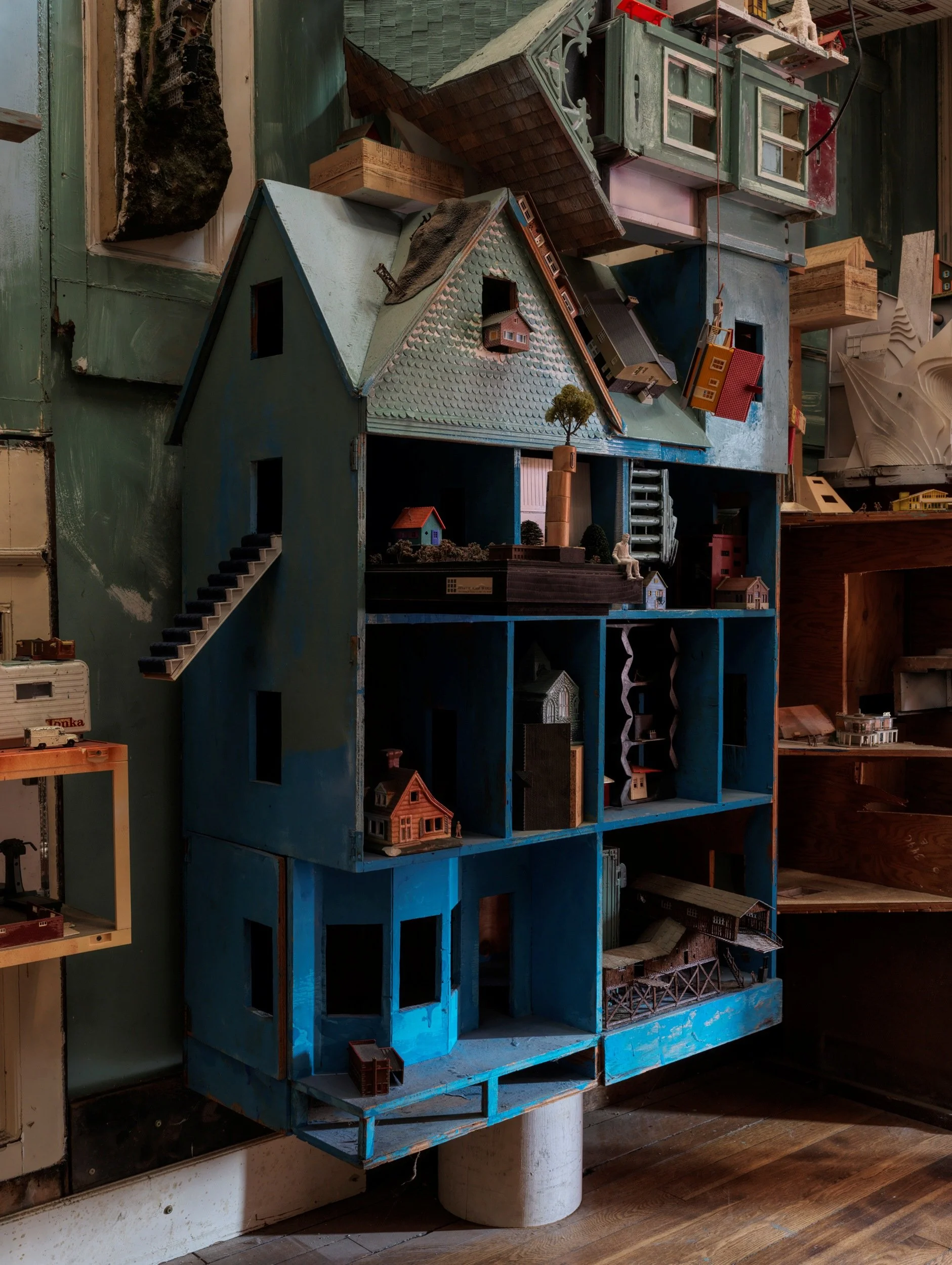 A detailed miniature model of a multi-story house with various other small buildings and trees inside a blue wooden display case.