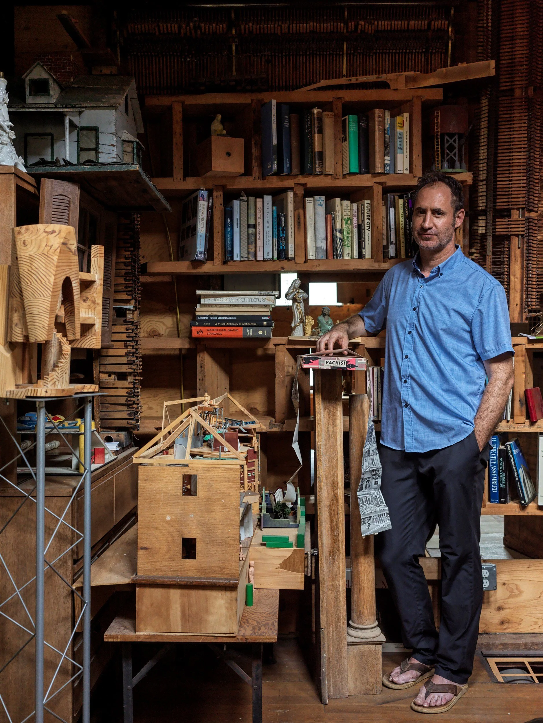 A man in a blue shirt and black pants stands in front of wooden shelves filled with books and architectural models, with a background of wooden interior and decorative objects.