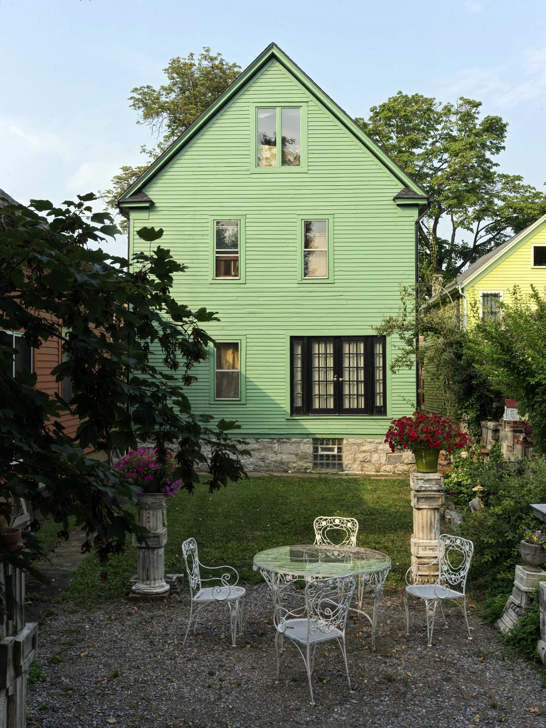 A garden backyard with a green wooden house, a white metal table, and chairs. There are flowering plants on stone pedestals and trees in the background.