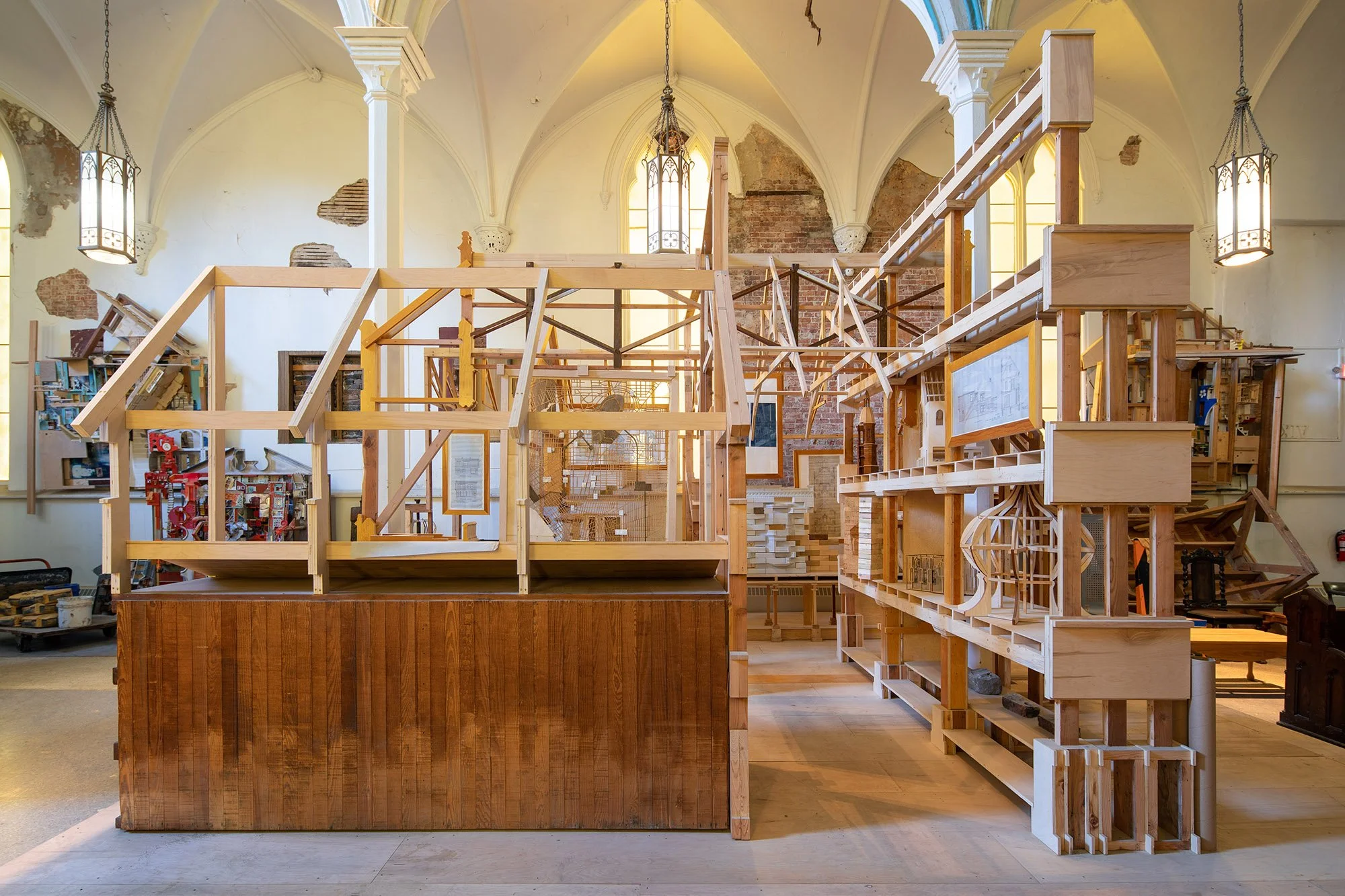 Interior of a church or old building undergoing construction, with wooden framework, scaffolding, high arched windows, brick walls, and hanging lantern lights.