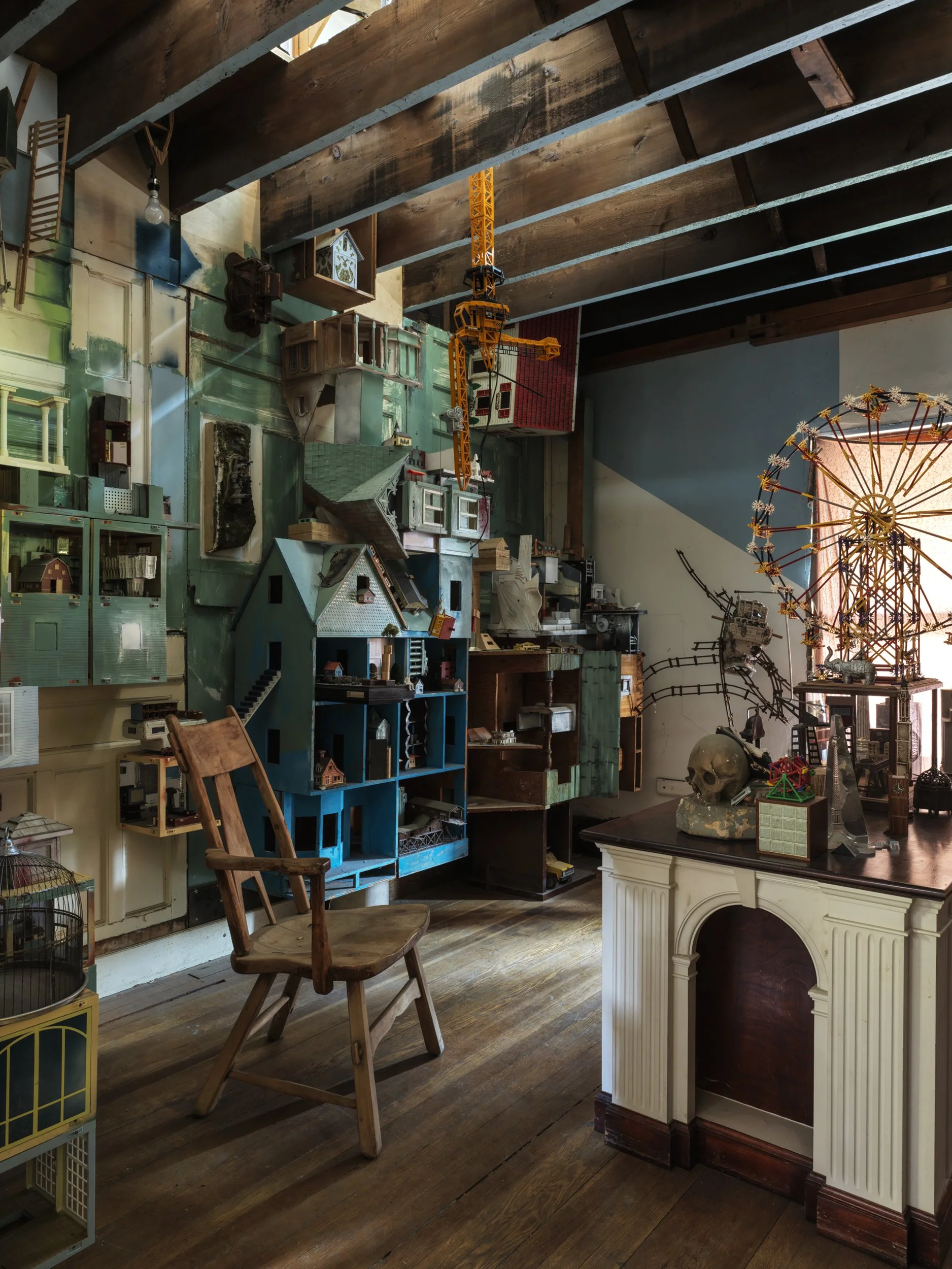 Interior of a room with wooden floor and ceiling, filled with miniature houses, a blue shelf with model houses, a wooden chair, a white fireplace mantel, and a ferris wheel decoration.