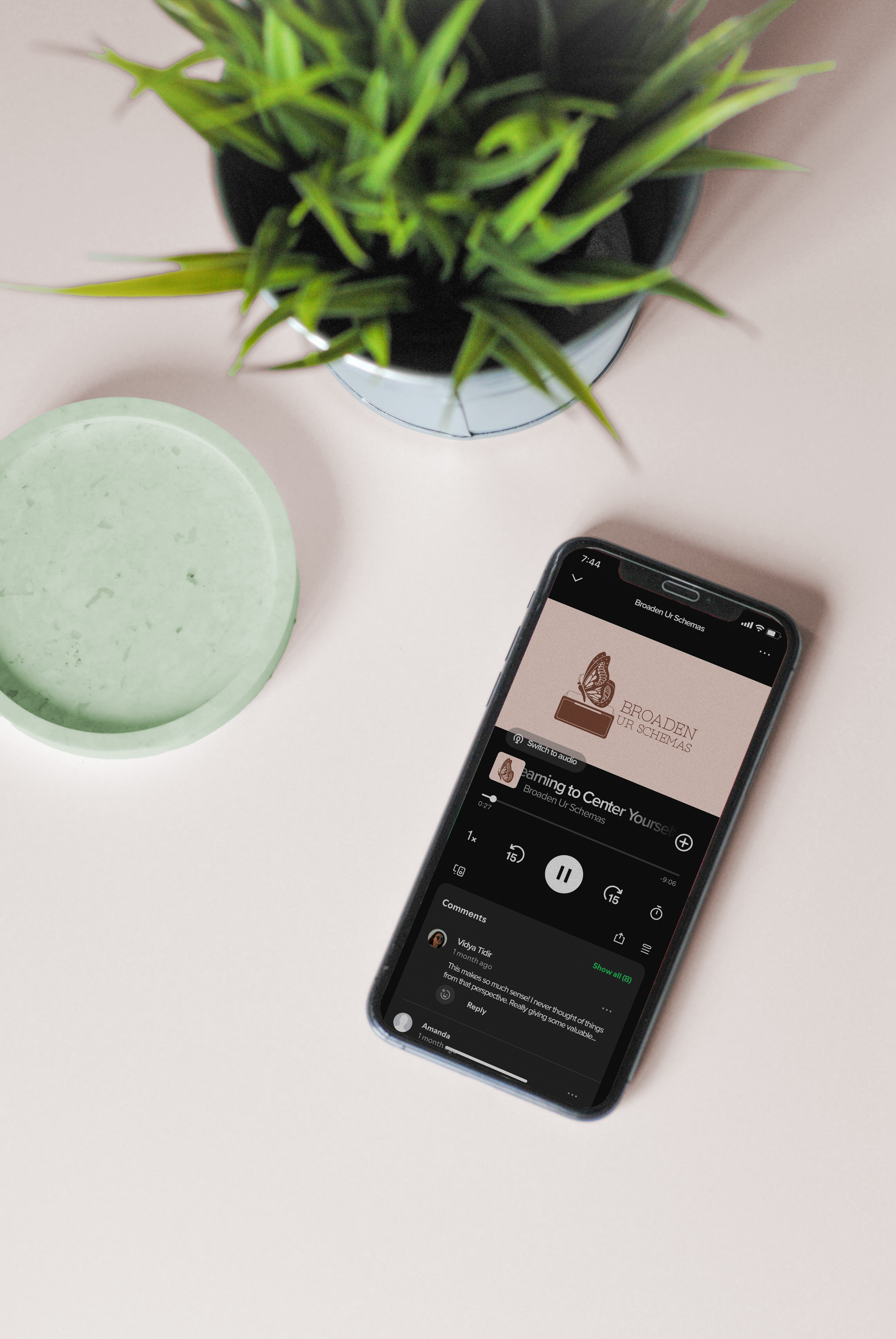 A smartphone on a white table displaying a podcast titled 'Learning to Center Yourself' by Broaden Ur Schemas, beside a potted green plant and a round green candle.