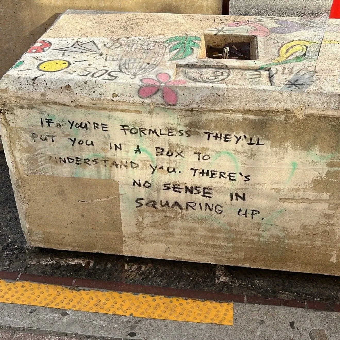 A concrete box with colorful graffiti and a quote painted on it, located on a sidewalk. The quote reads, 'If you're formless they'll put you in a box to understand you. There's no sense in squaring up.'
