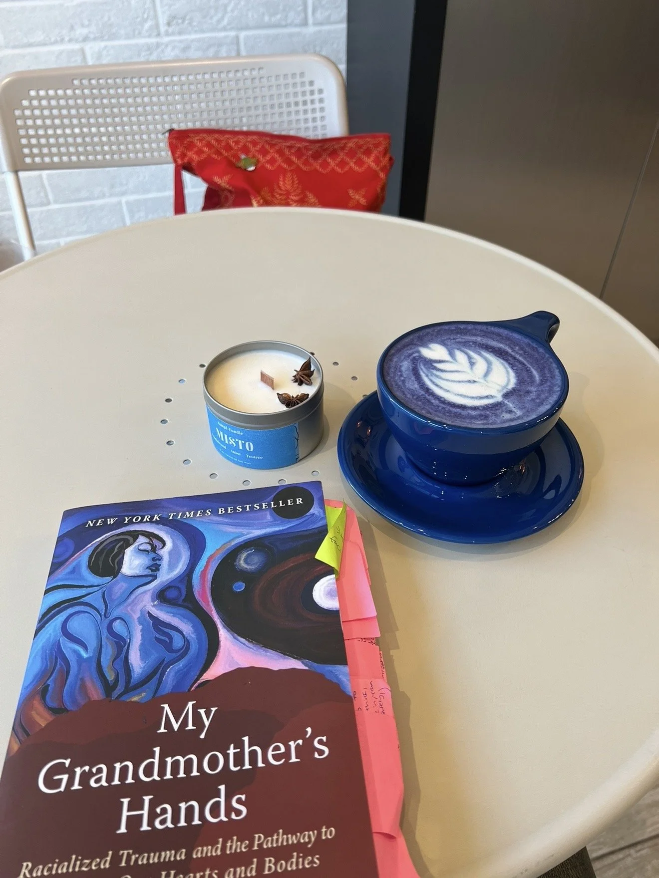 A white round table holding a blue cup of purple latte with latte art, a blue candle with star anise, and a book titled "My Grandmother's Hands." In the background, there is a white chair with a red pillow and a gray wall with a brick pattern.
