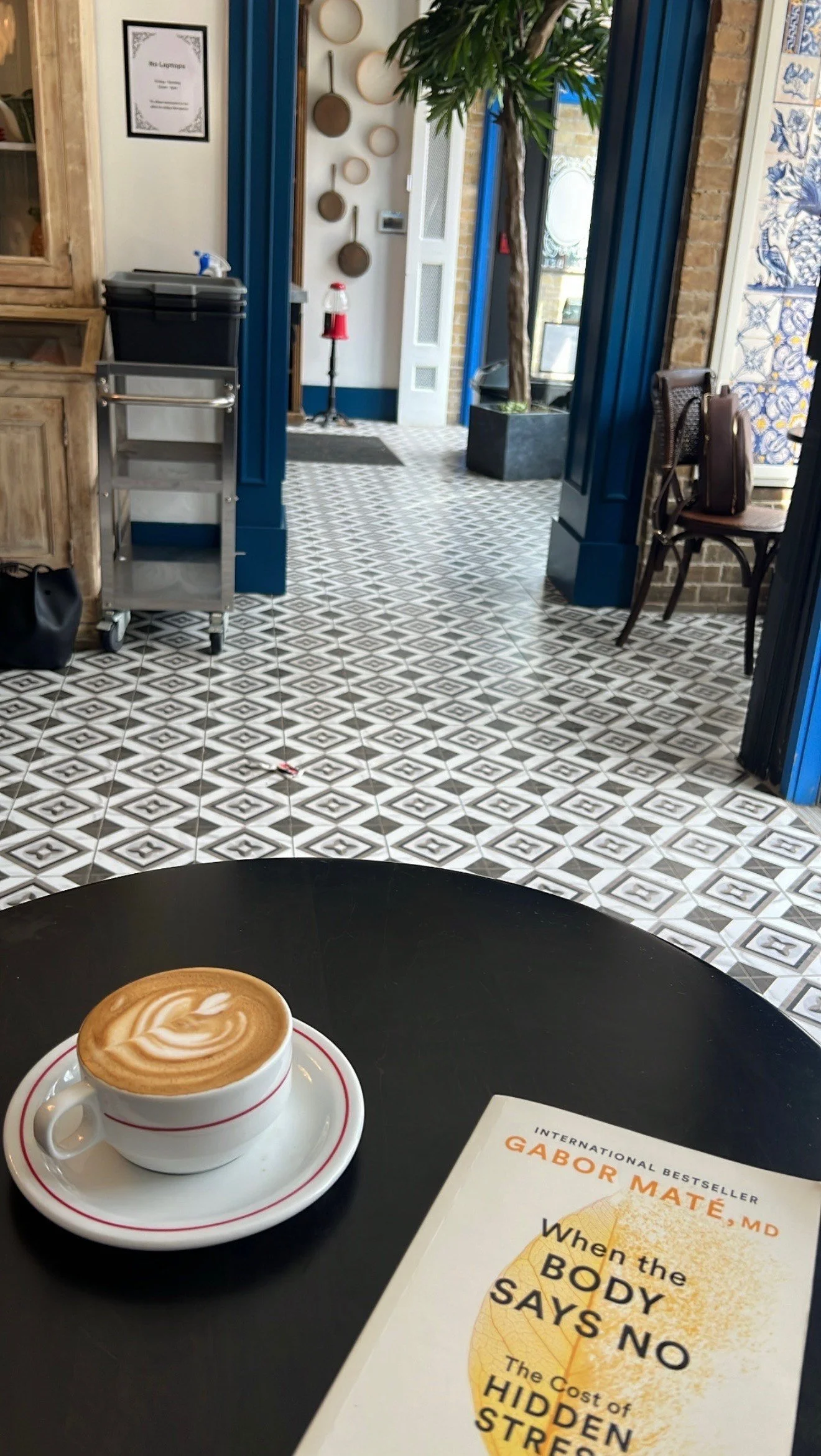 A cup of latte with foam art on a black table, a book titled 'When the Body Says No' by Gabor Maté, and a cozy cafe interior with patterned floor tiles, blue trim, and decorative wall hanging.