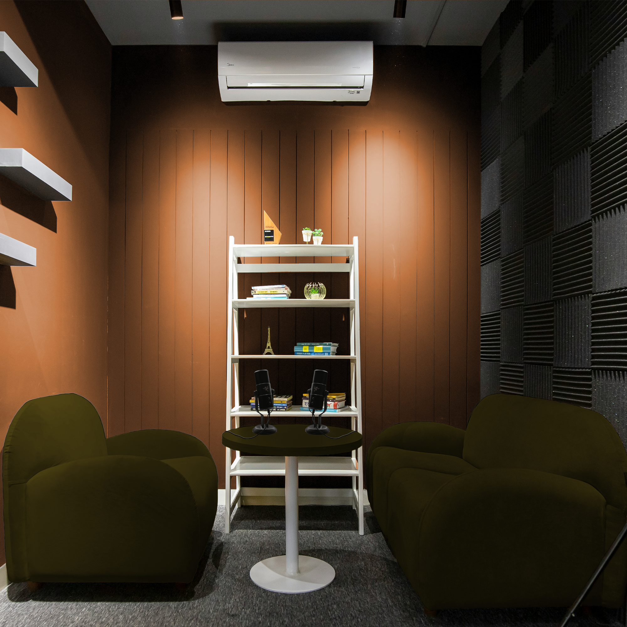 Podcast recording studio with two green armchairs facing each other, a small round table with two microphones, a white shelf with books and decor, and a brick wall with soundproof panels.