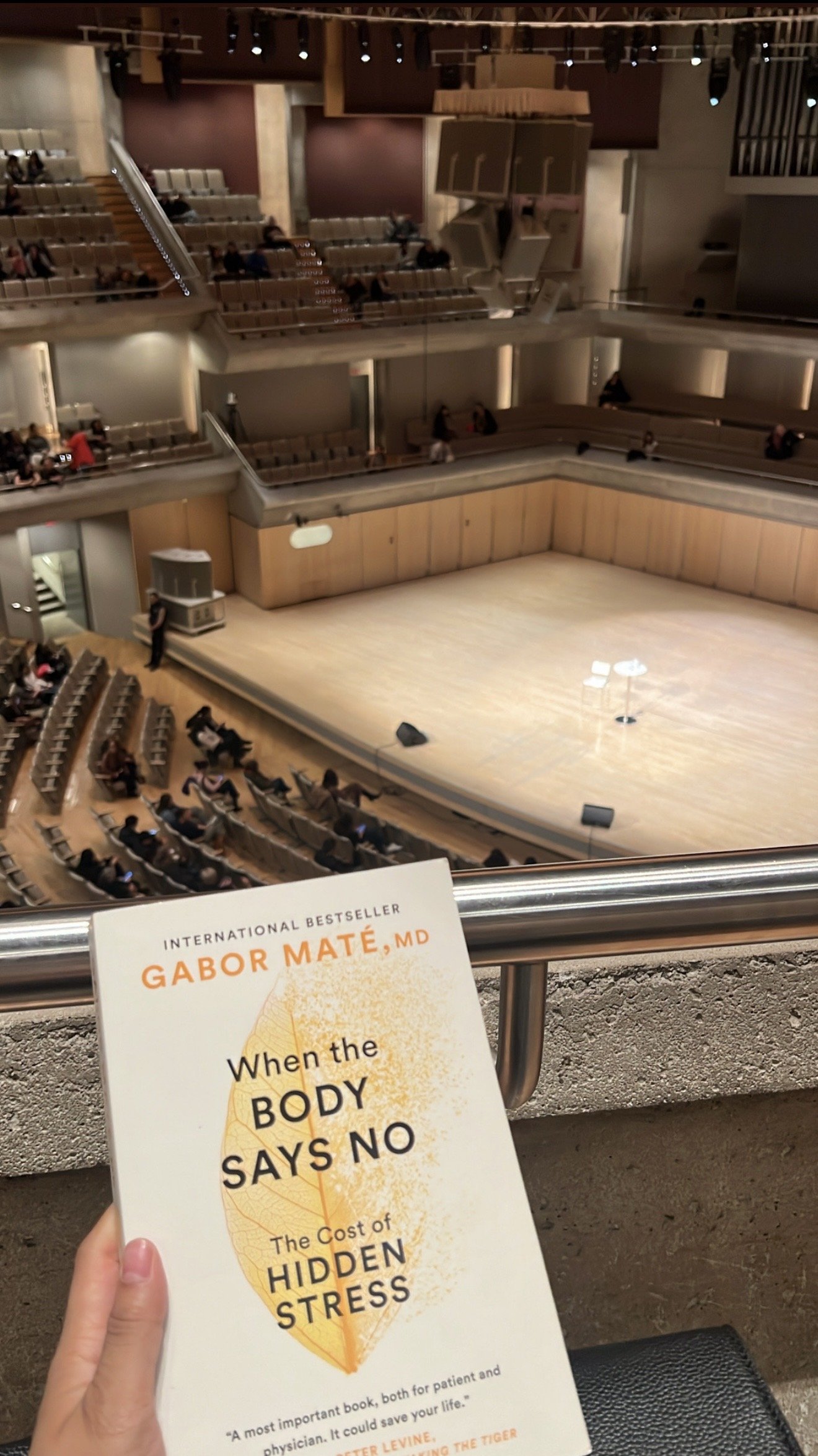An empty theater stage with chairs and microphones, viewed from balcony seating. In the foreground, a person holds a book titled 'When the Body Says No: The Cost of Hidden Stress' by Gabor Maté, MD.
