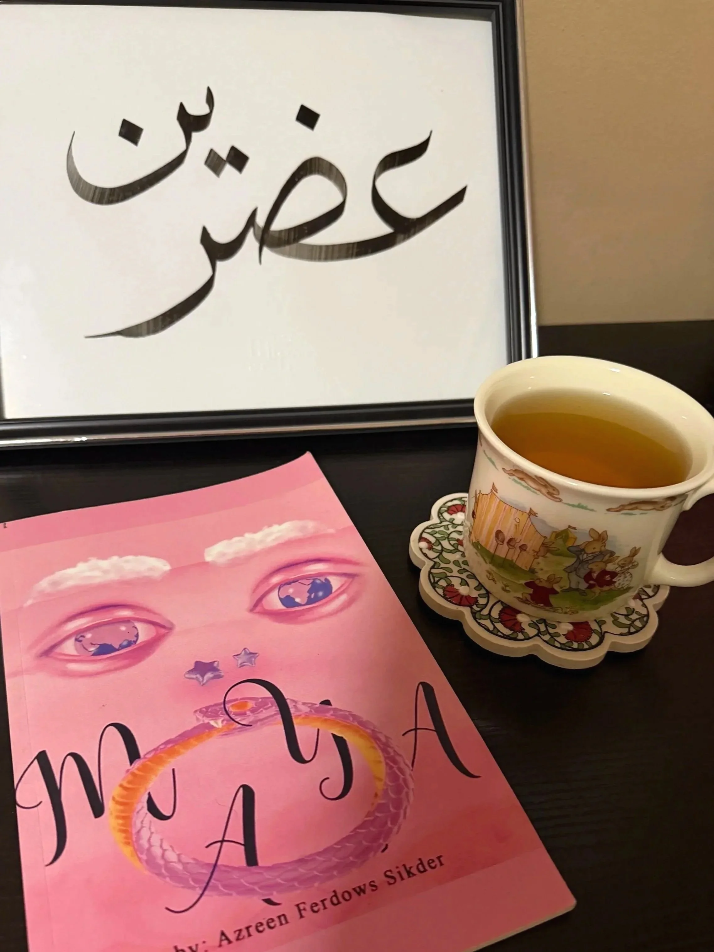 A framed piece of art with Arabic calligraphy in black ink, a pink book titled "Maya" with an illustration of a girl and a snake, and a cup of tea in a decorative mug on a black table.