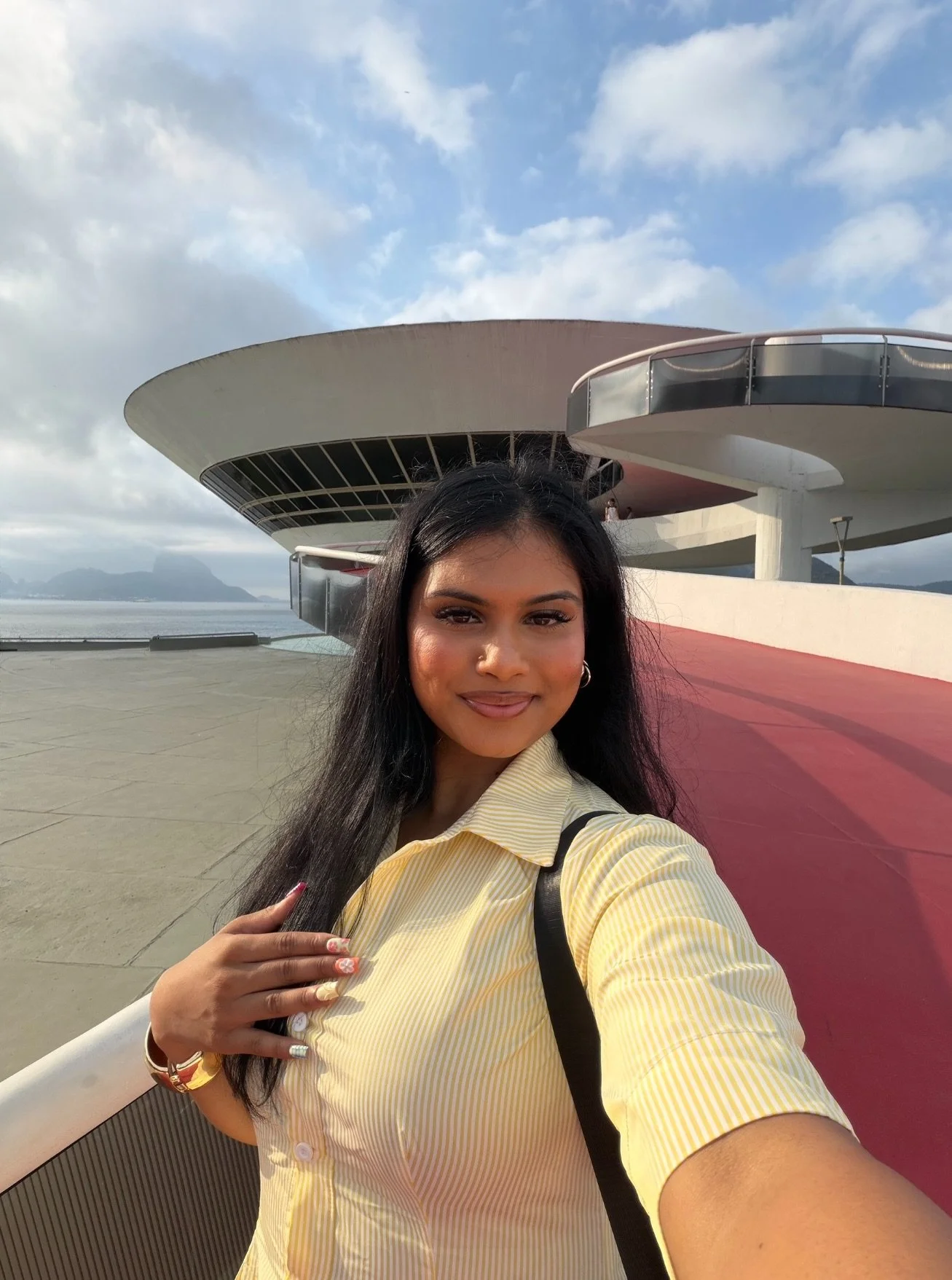 A woman taking a selfie outdoors near a modern building with a circular architectural design under a partly cloudy sky.