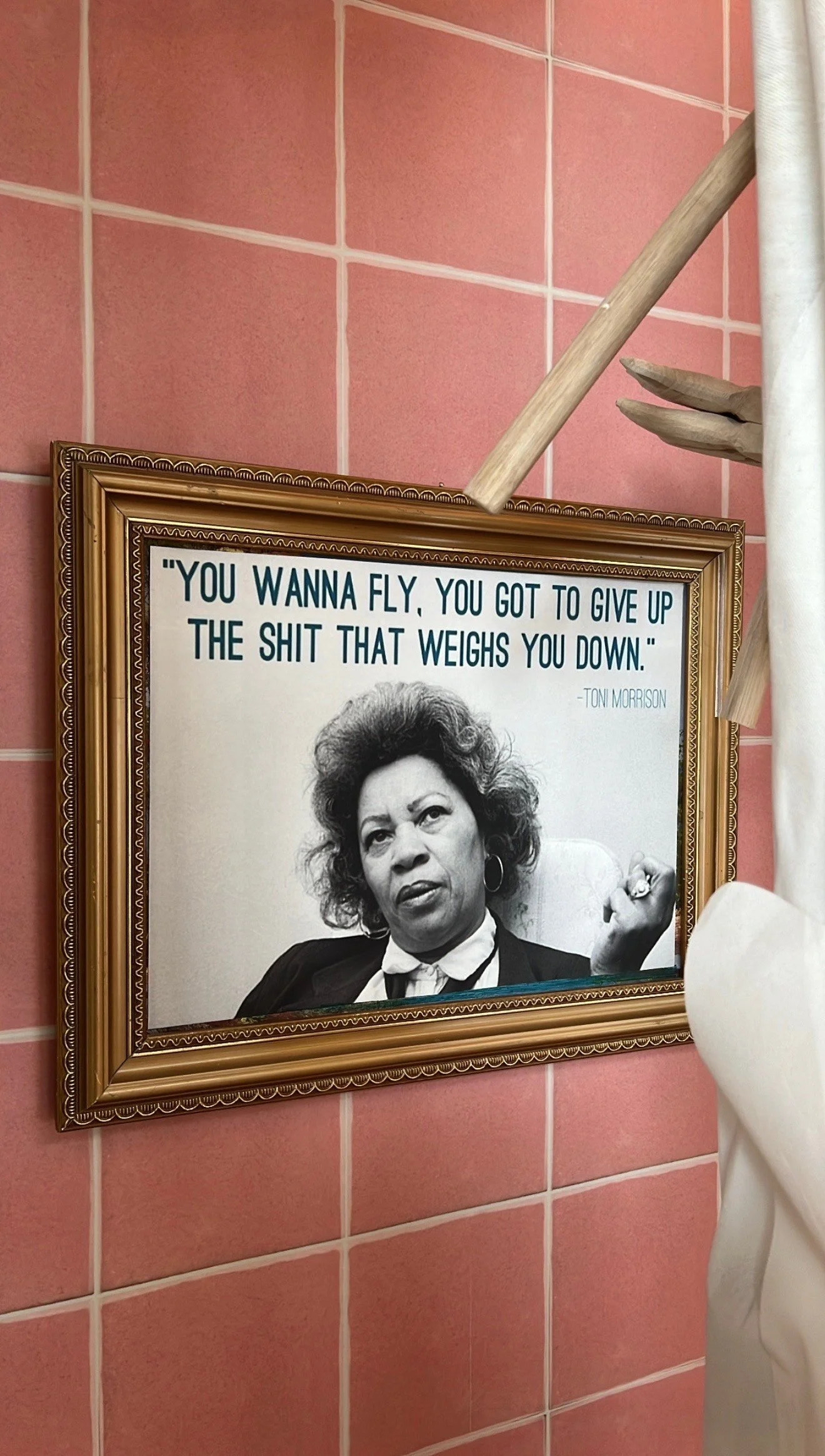 A framed quote on a pink tiled wall features a black-and-white photo of a woman with curly hair, wearing a dark blazer and earrings. The quote reads: "You wanna fly, you got to give up the shit that weighs you down." - Toni Morrison.