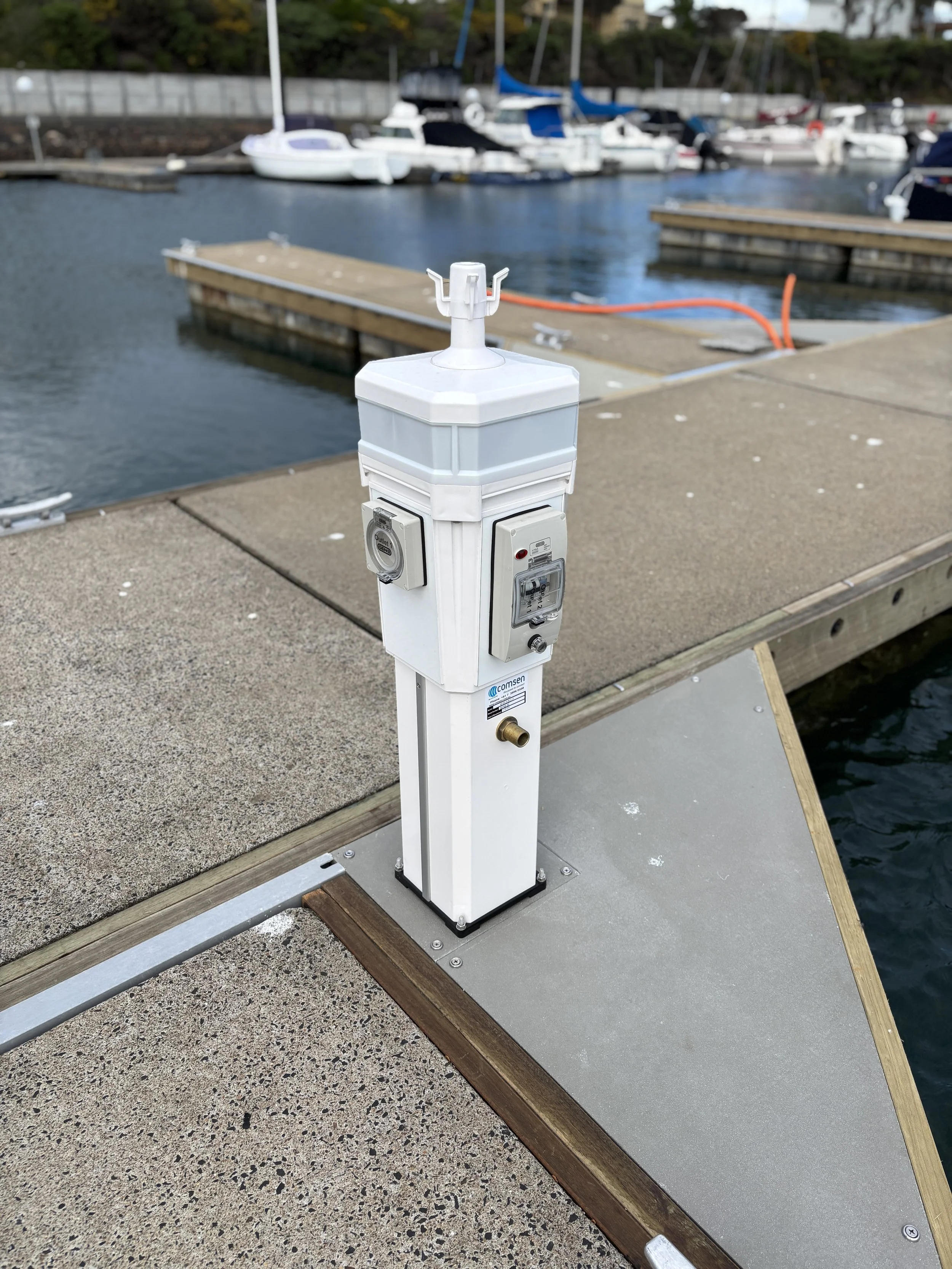 A dockside water meter and utility box with pipes and cables, near boats in a marina.