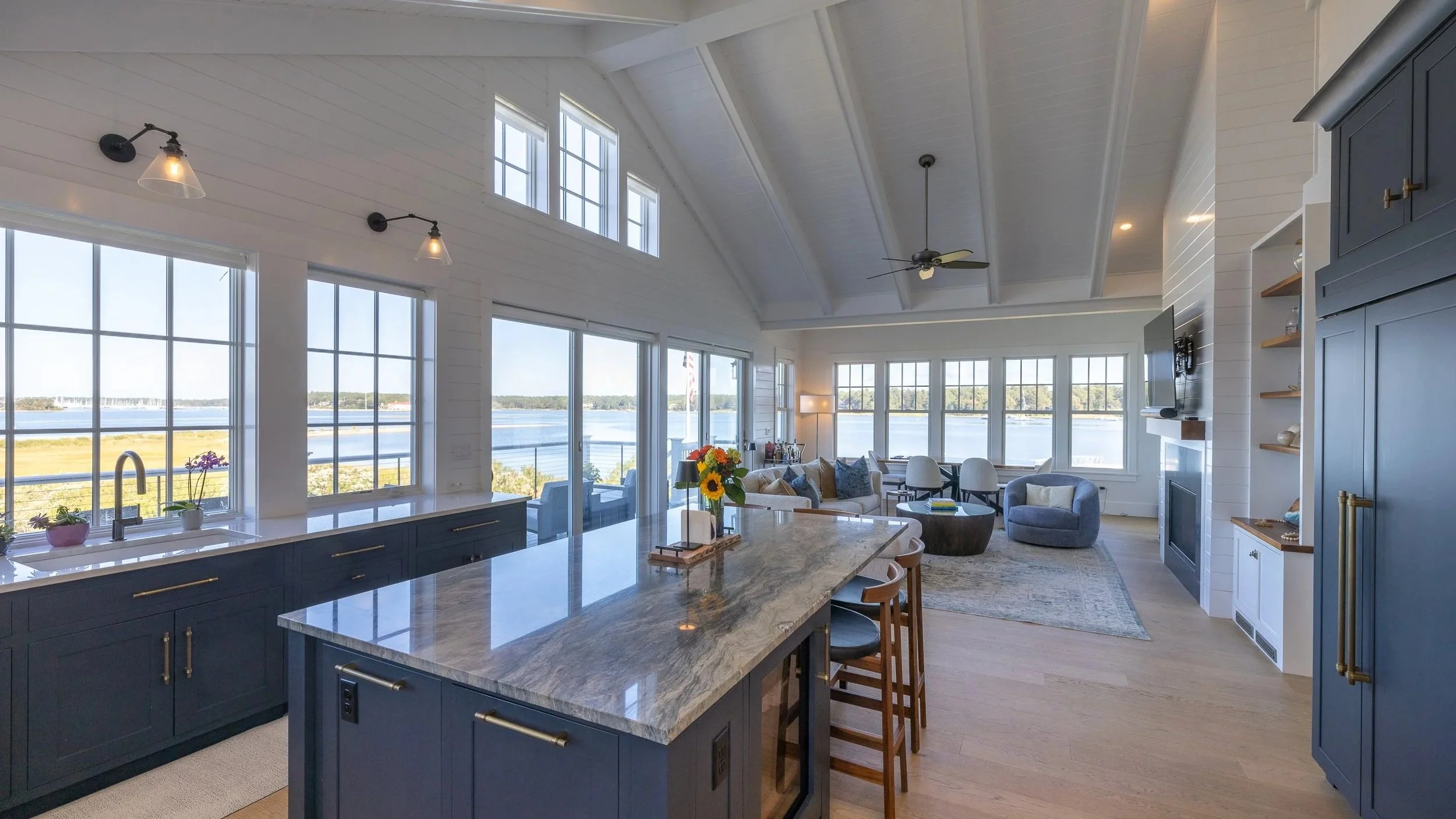 Open-concept living room and kitchen with large windows overlooking water, blue cabinetry, marble island, and coastal decor.