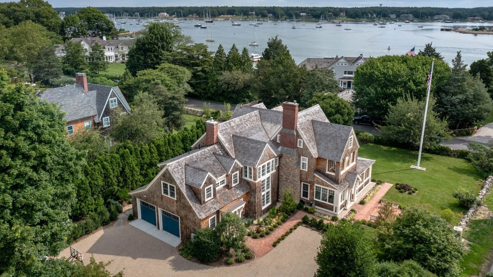 A large residential house with multiple roof angles and brick chimneys situated near a body of water with boats, surrounded by green trees and lawns, with an American flag flying on a flagpole.