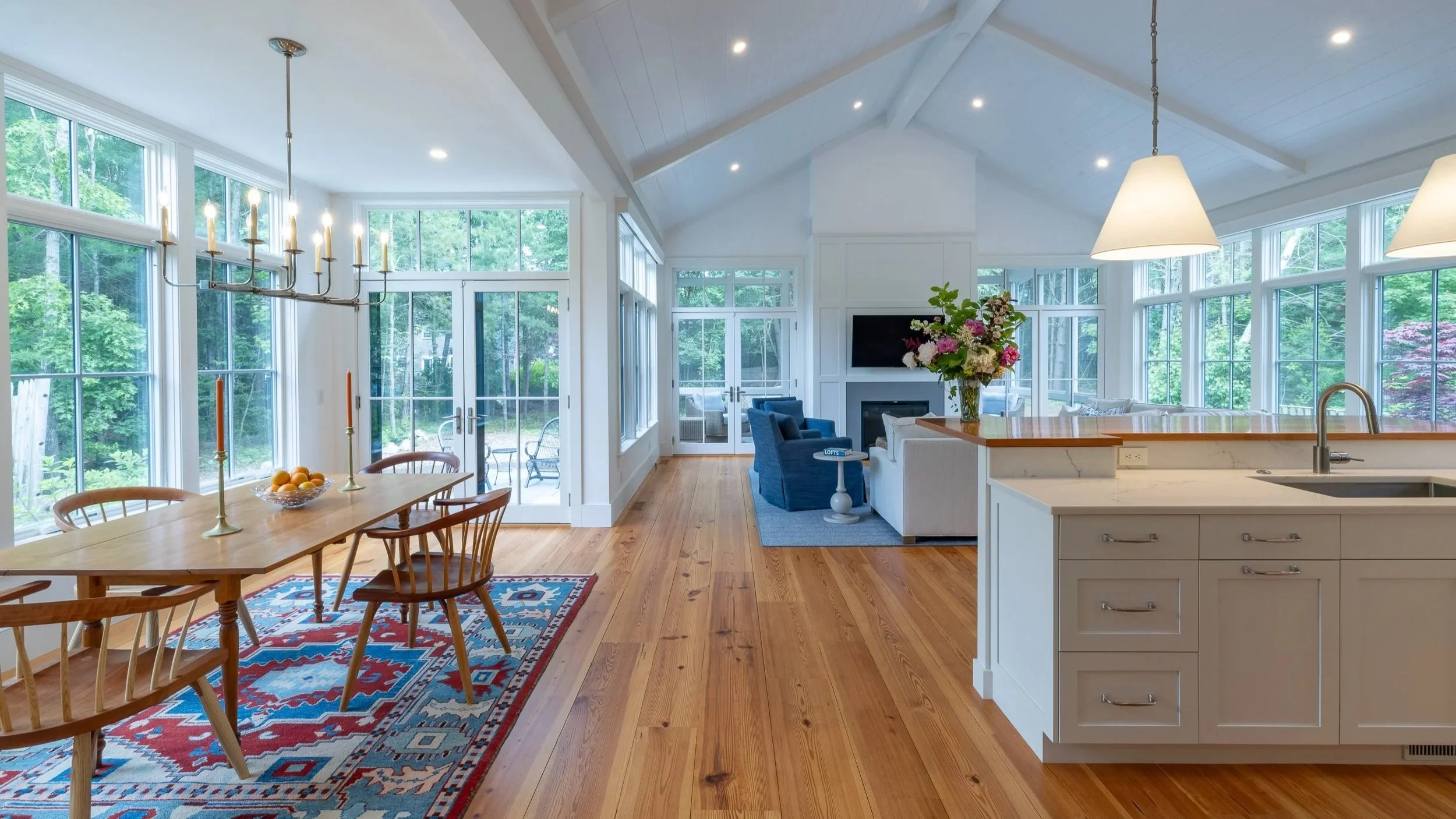Bright open-concept living and dining area with large windows, wood floors, a dining table with chairs, a chandelier, a kitchen island with pendant lights, and a cozy sitting area with sofas and a fireplace.