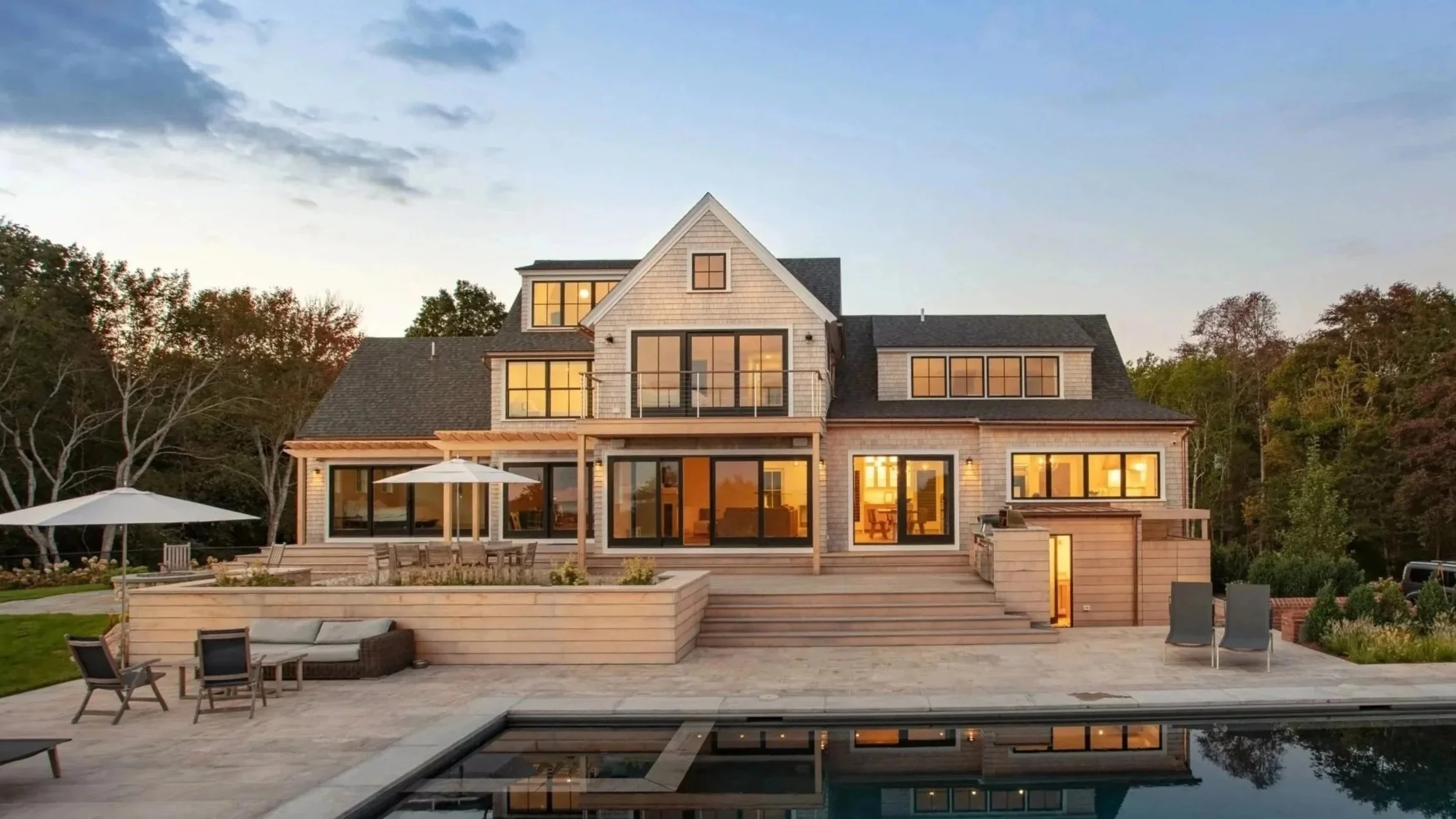 A modern, multi-story house with large glass windows and a backyard at sunset, featuring outdoor furniture, umbrellas, and a swimming pool in the foreground.