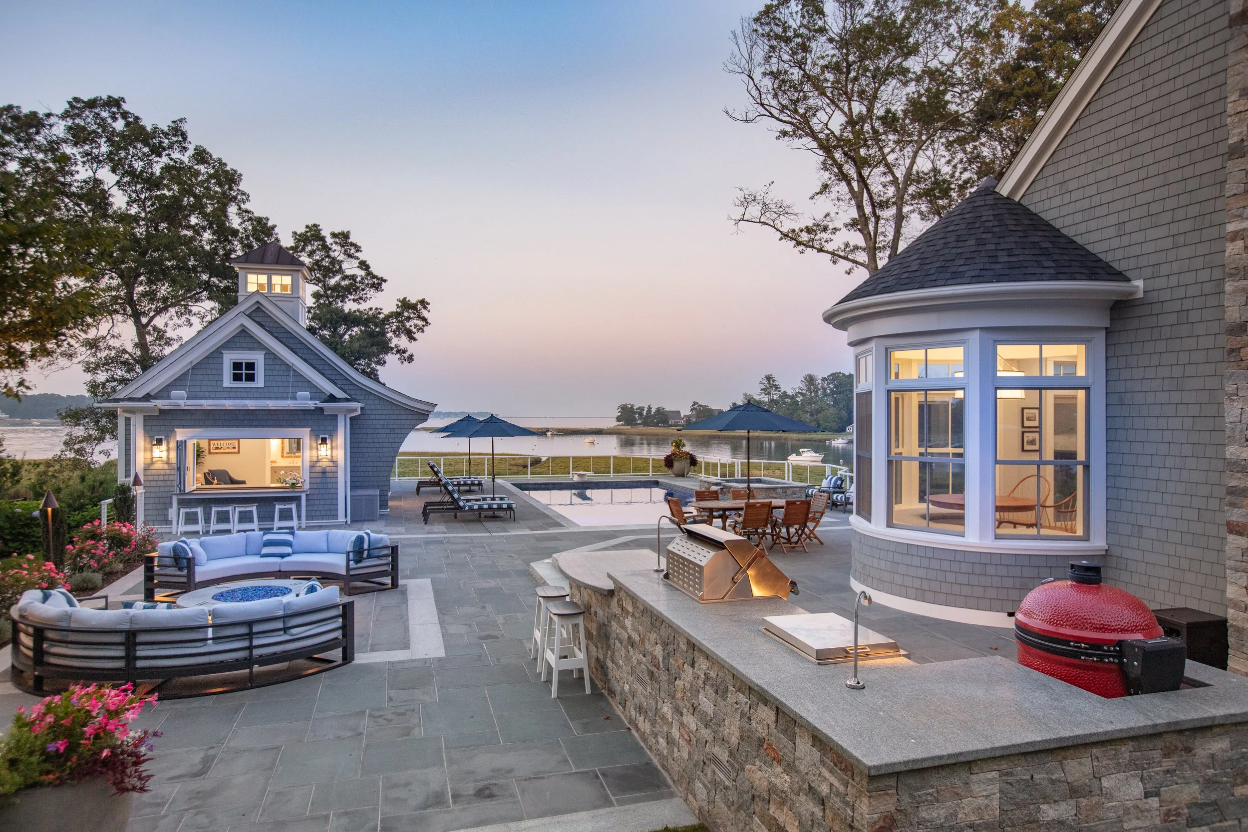 Outdoor patio with seating area, a bar, a pool with umbrellas, and a view of a lake at sunset.