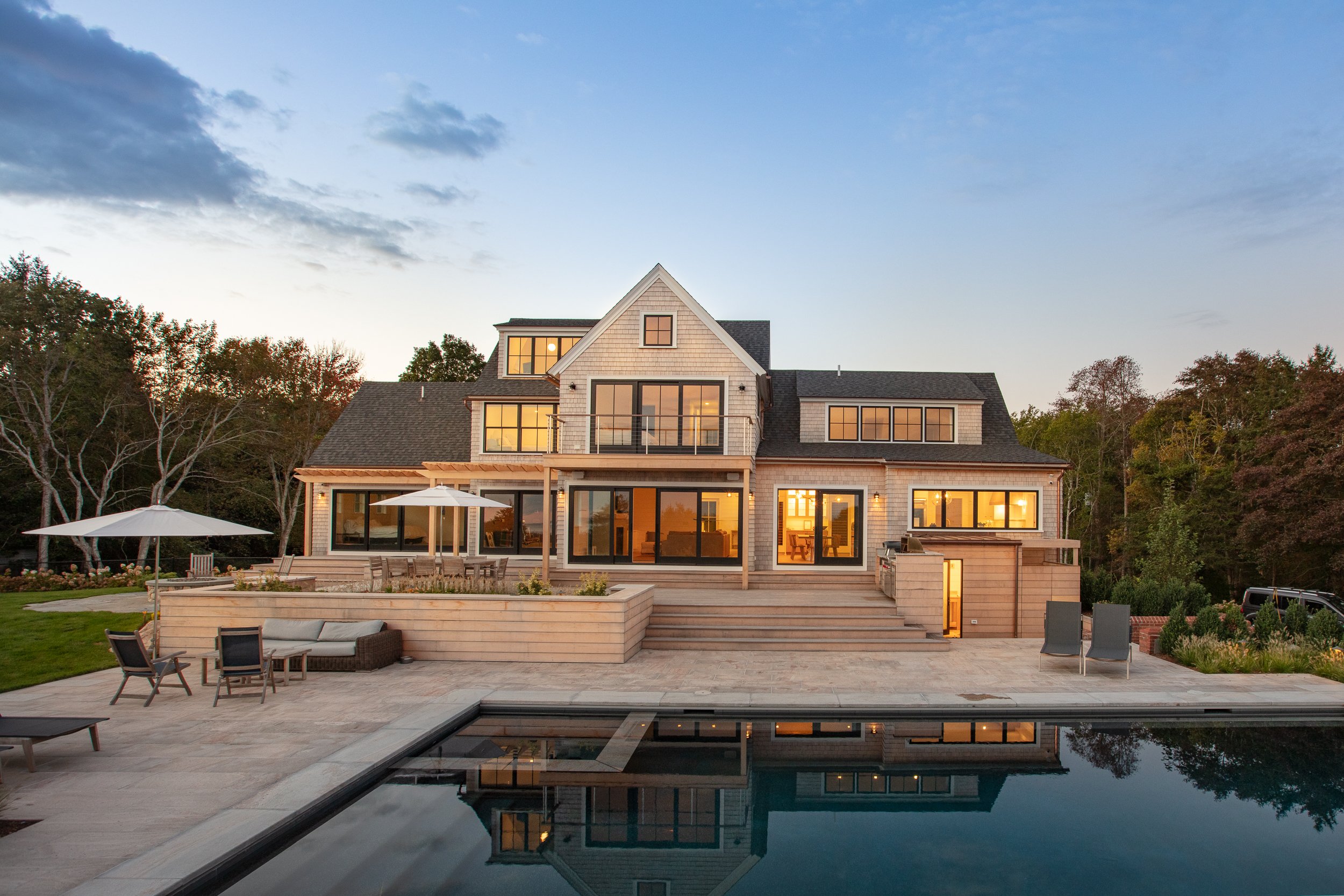 A modern multi-story house with large windows and a backyard with a swimming pool, outdoor chairs, umbrellas, and a deck at sunset, surrounded by trees.