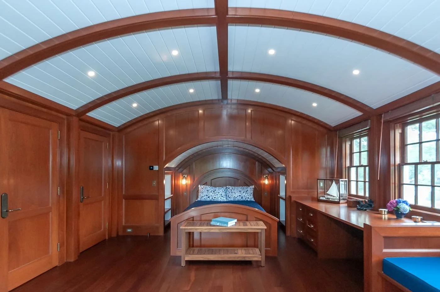 Bedroom with wooden paneling, arched ceiling, and large windows; there is a bed with a wooden headboard, a bench at the foot of the bed, and a long wooden dresser with decorative items.