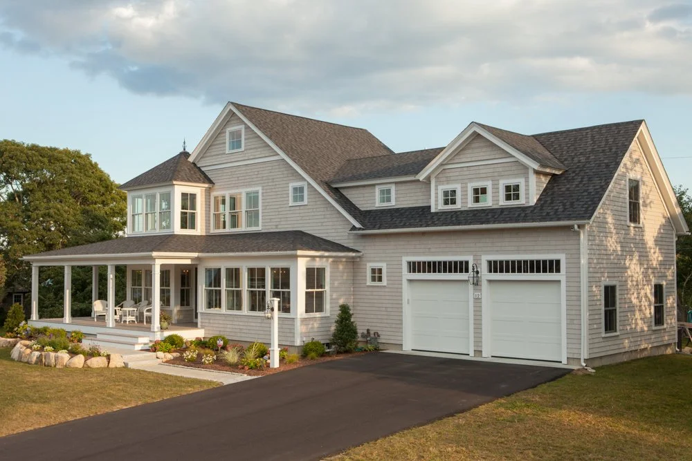A large two-story house with light-colored siding, front porch with seating, attached two-car garage, and a landscaped yard with flowers and shrubs.