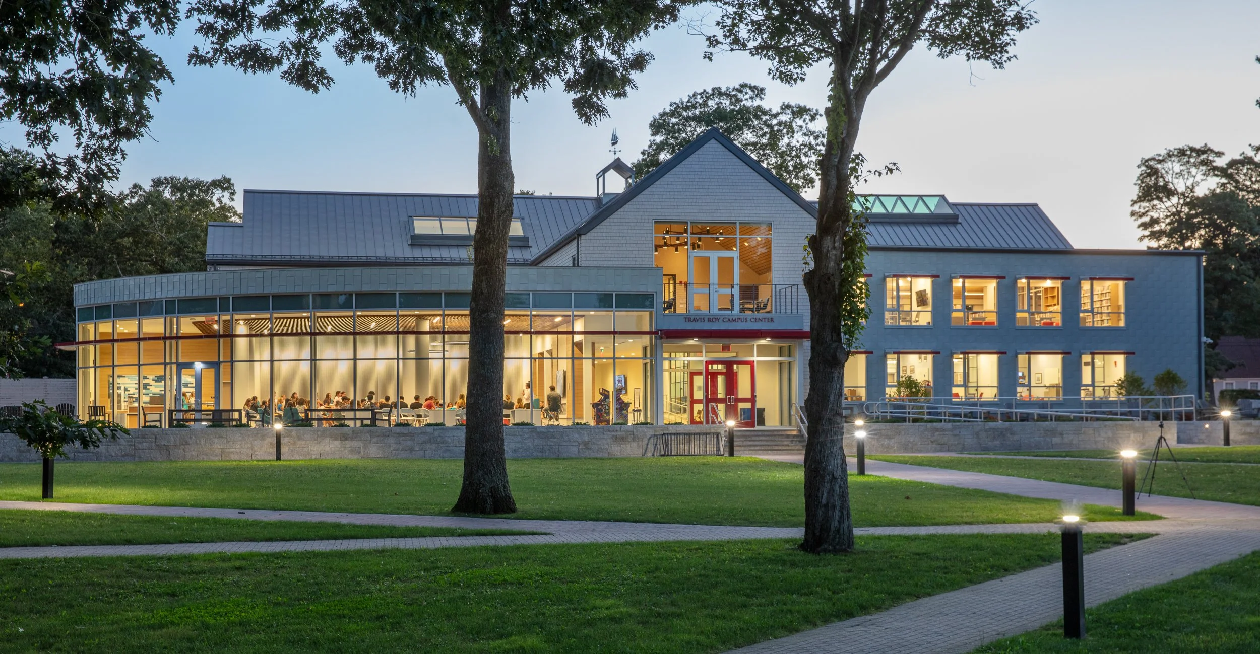 A modern campus center building with large glass windows, illuminated from inside, surrounded by a well-maintained grassy area and walking paths, with trees and outdoor lights, during evening hours.