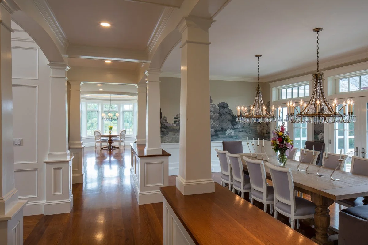 Photo of a spacious dining room with a long wooden table set for a meal, adorned with a flower arrangement and candle holders. Two ornate chandeliers hang above the table, and large windows let in natural light. The background shows a wall mural of a lakeside scene.