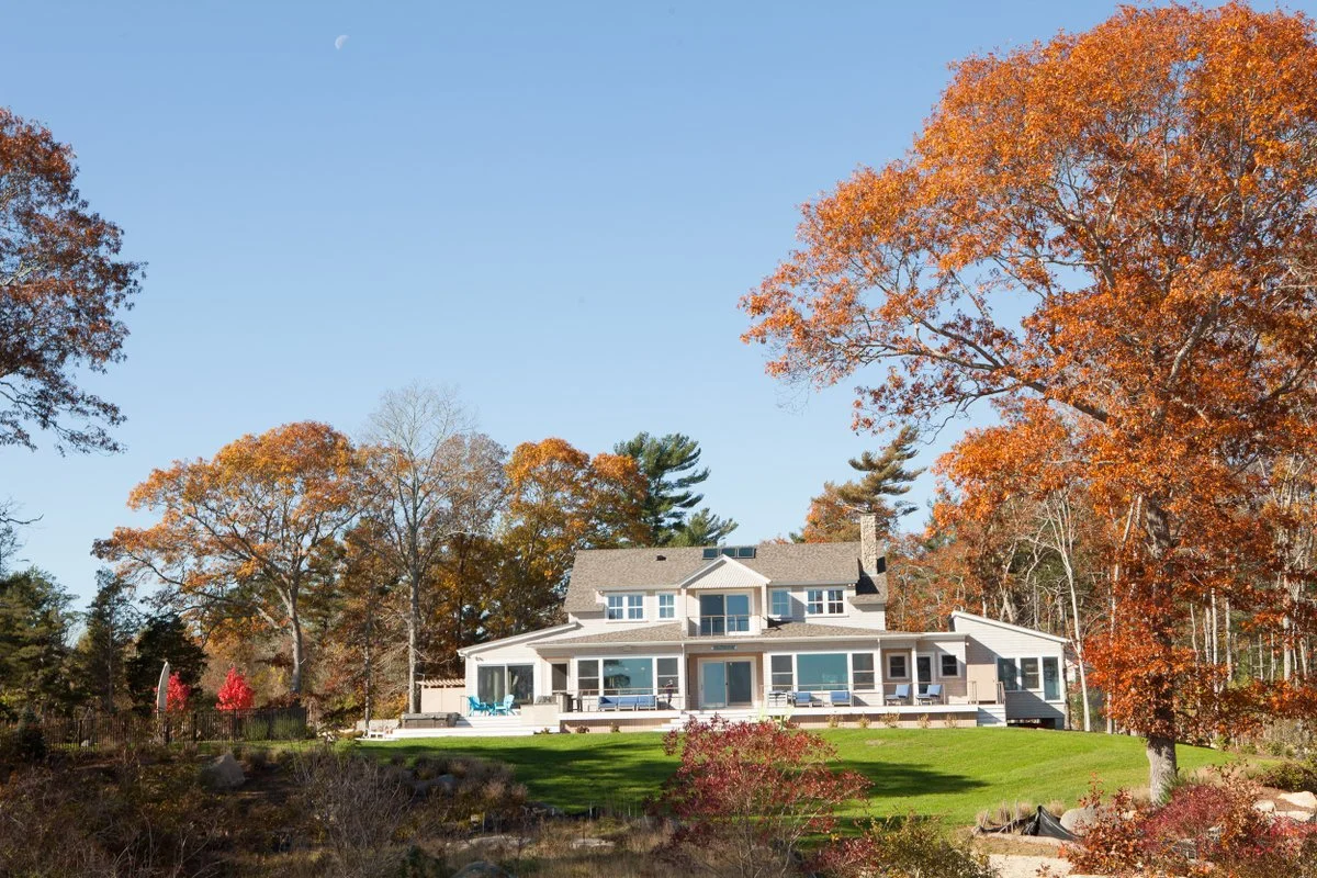 Large modern house with a porch and balcony, surrounded by autumn trees with orange leaves, under a clear blue sky.