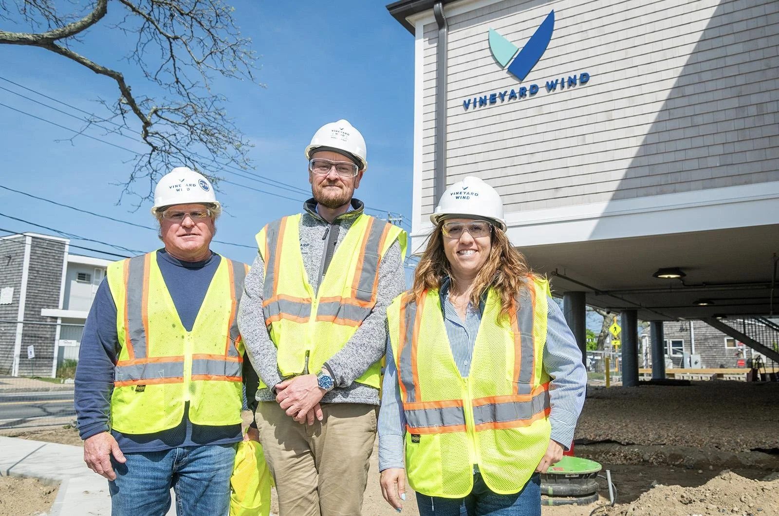 Three construction workers in yellow safety vests and white helmets standing outside a building with a "Vineyard Wind" sign, under a blue sky.