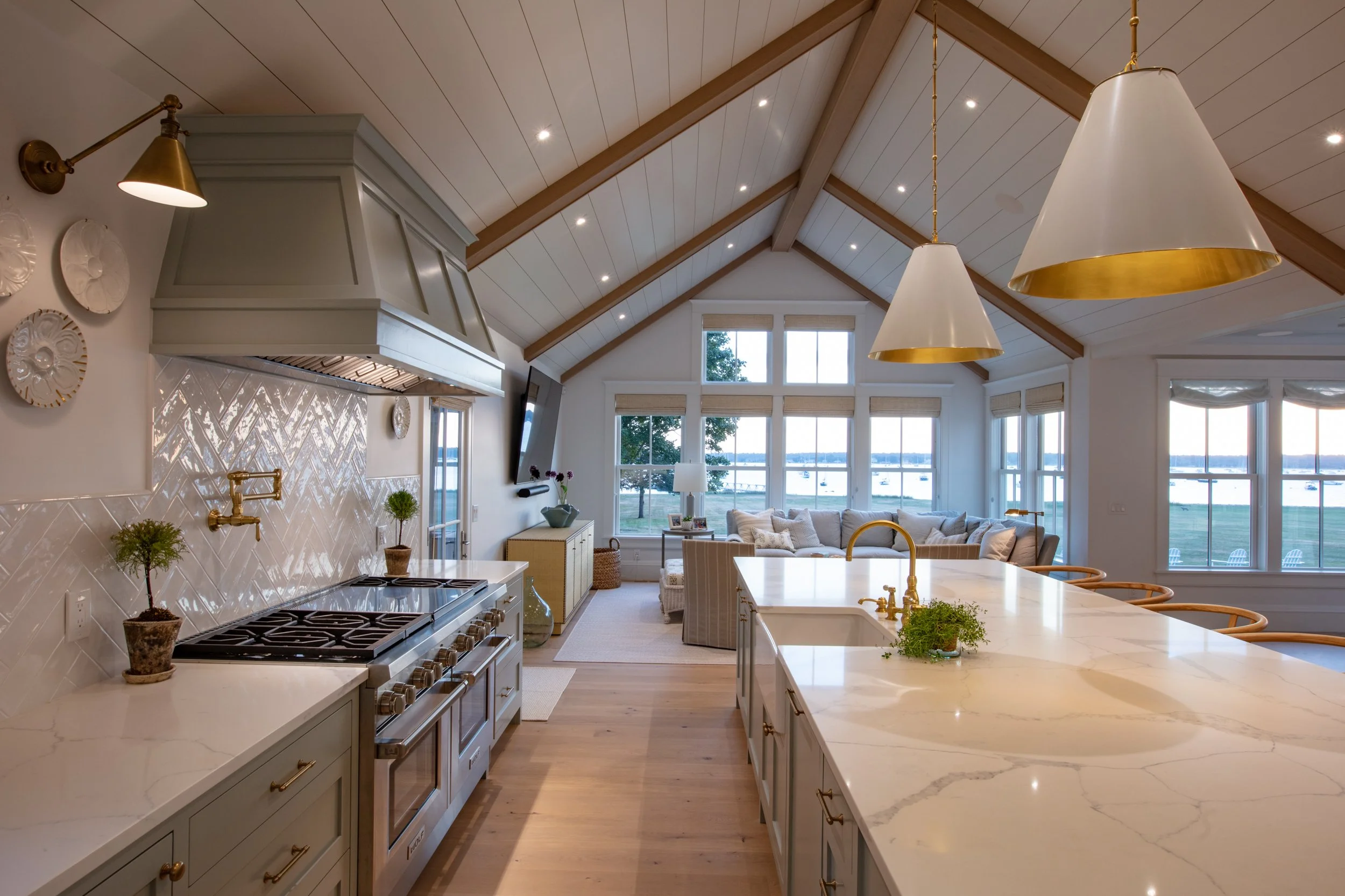 Bright, open-concept kitchen and living room with vaulted ceiling, hanging pendant lights, large windows overlooking a lake, and a kitchen island with a marble countertop.