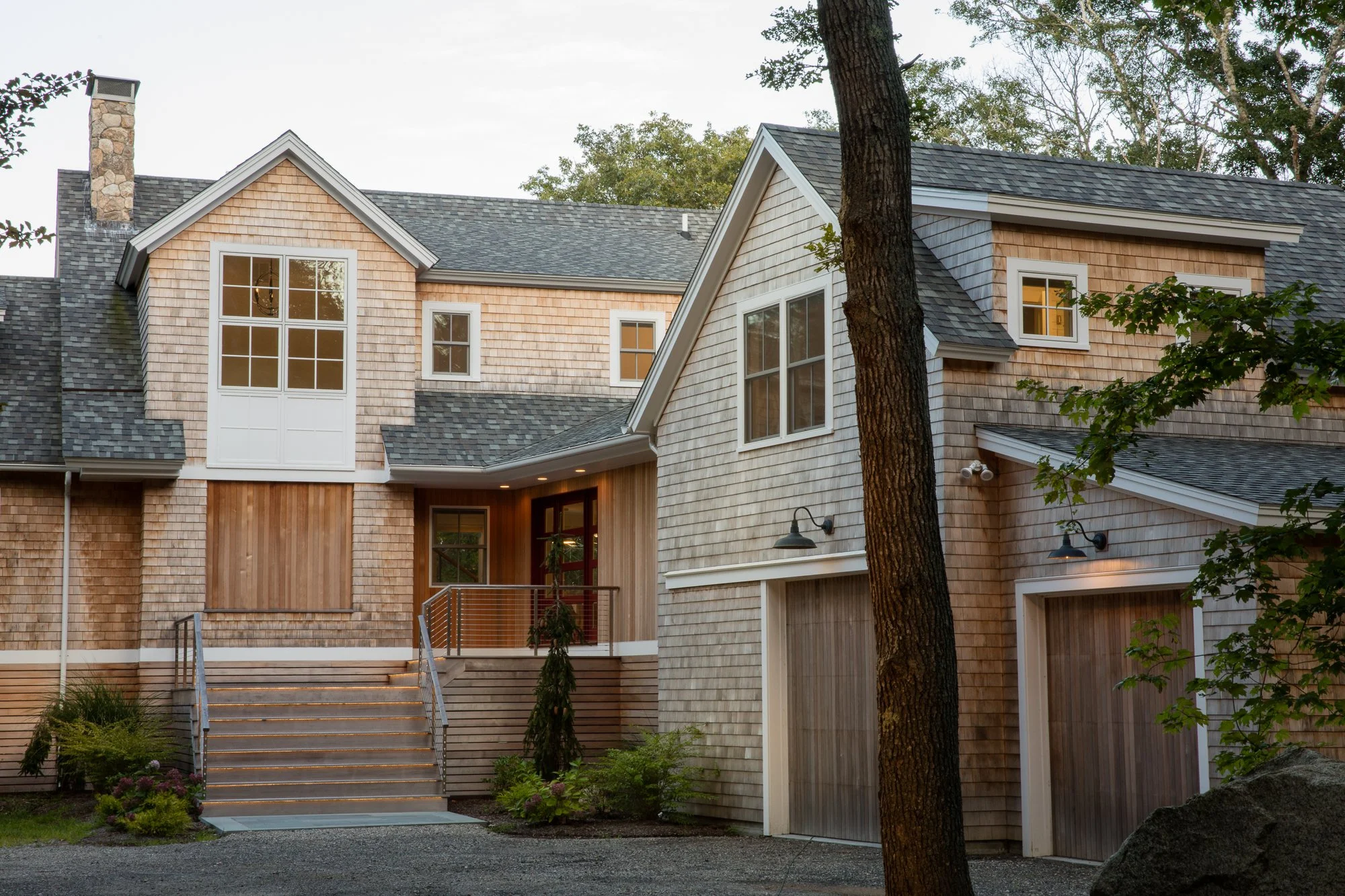A modern two-story house with wooden shingles, multiple windows, a front staircase, and outdoor lighting in a green yard.