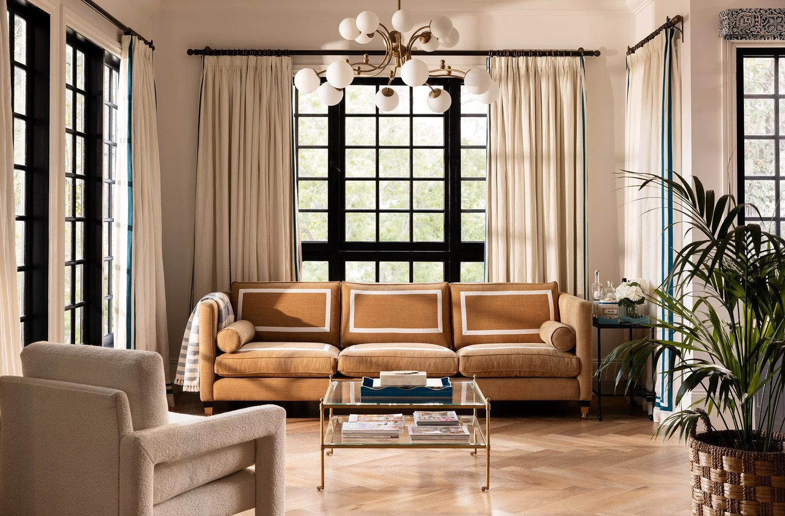 Living room with beige couch, cream armchair, glass coffee table, large windows with black frames, cream curtains, ceiling chandelier with white globes, green potted plant, side table with water bottles and flowers.