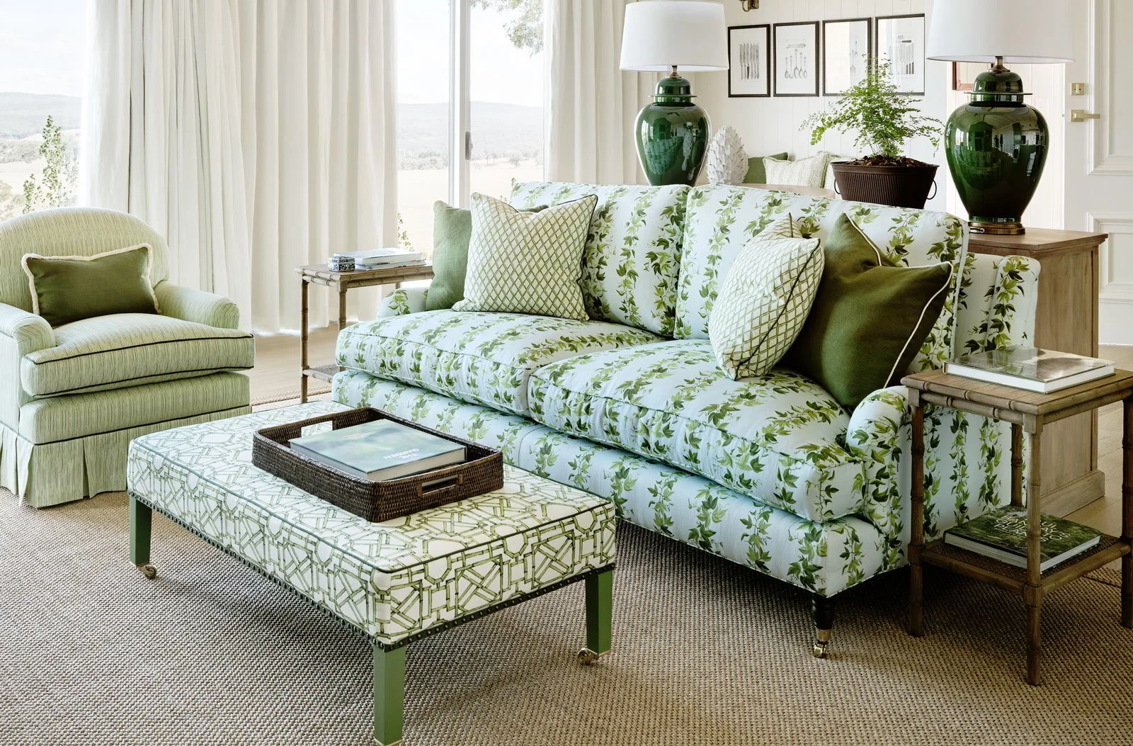 Living room with green and white floral sofa, matching throw pillows, green armchair, side tables with books, large green lamps, potted plant, and framed pictures on the wall, daylight coming through large window with cream curtains, carpeted floor.