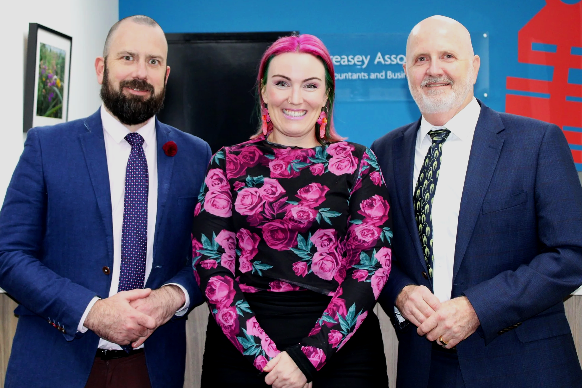 Three people standing inside an office, smiling at the camera. The person on the left is a man with a beard, wearing a blue suit, white shirt, and patterned tie. The person in the middle is a woman with pink and green hair, wearing a black dress with pink roses. The person on the right is a man with a white beard, wearing a dark suit, white shirt, and a patterned tie.