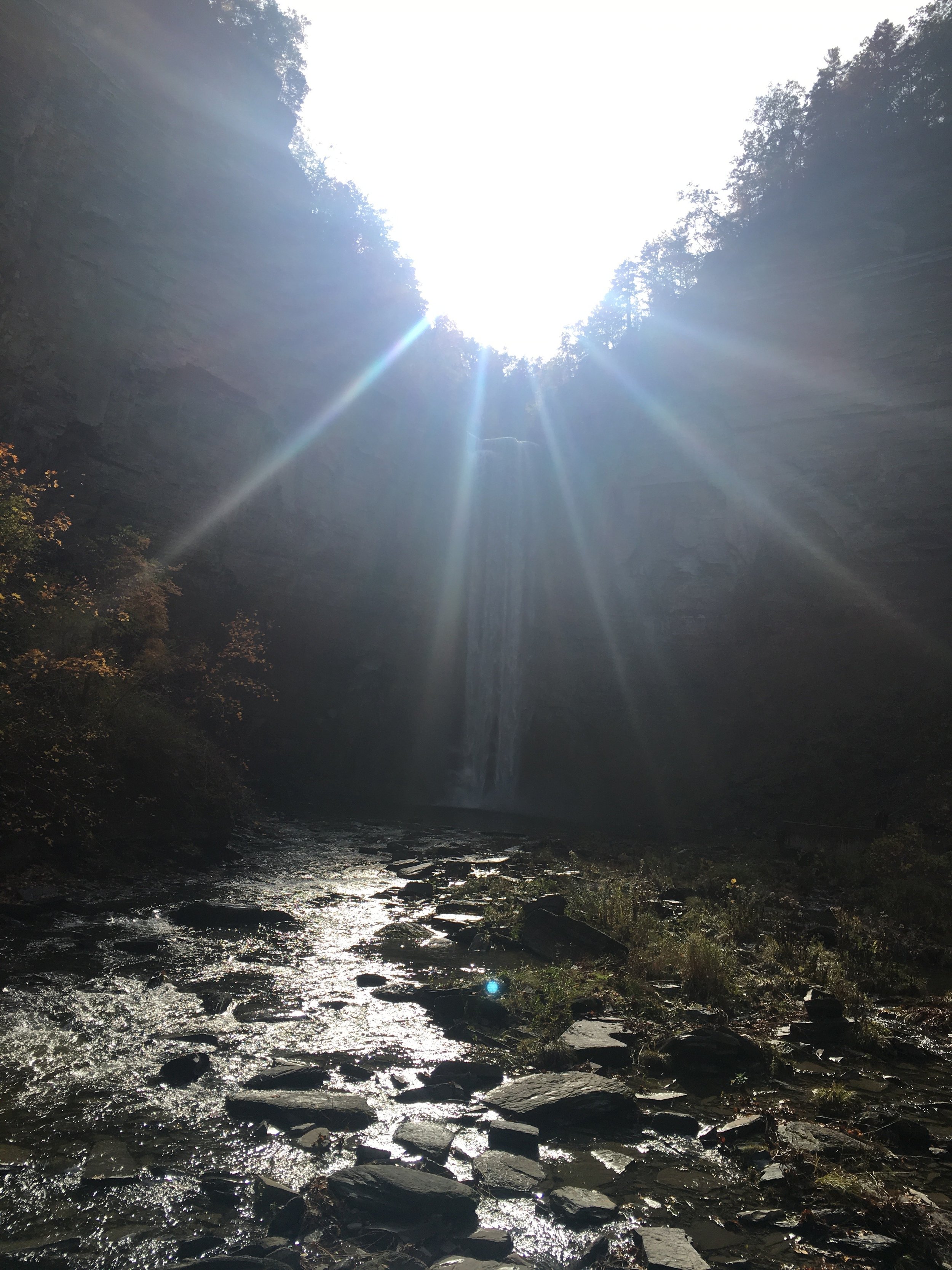 Sunlight shining through the top of a canyon, illuminating a waterfall and a rocky river below.