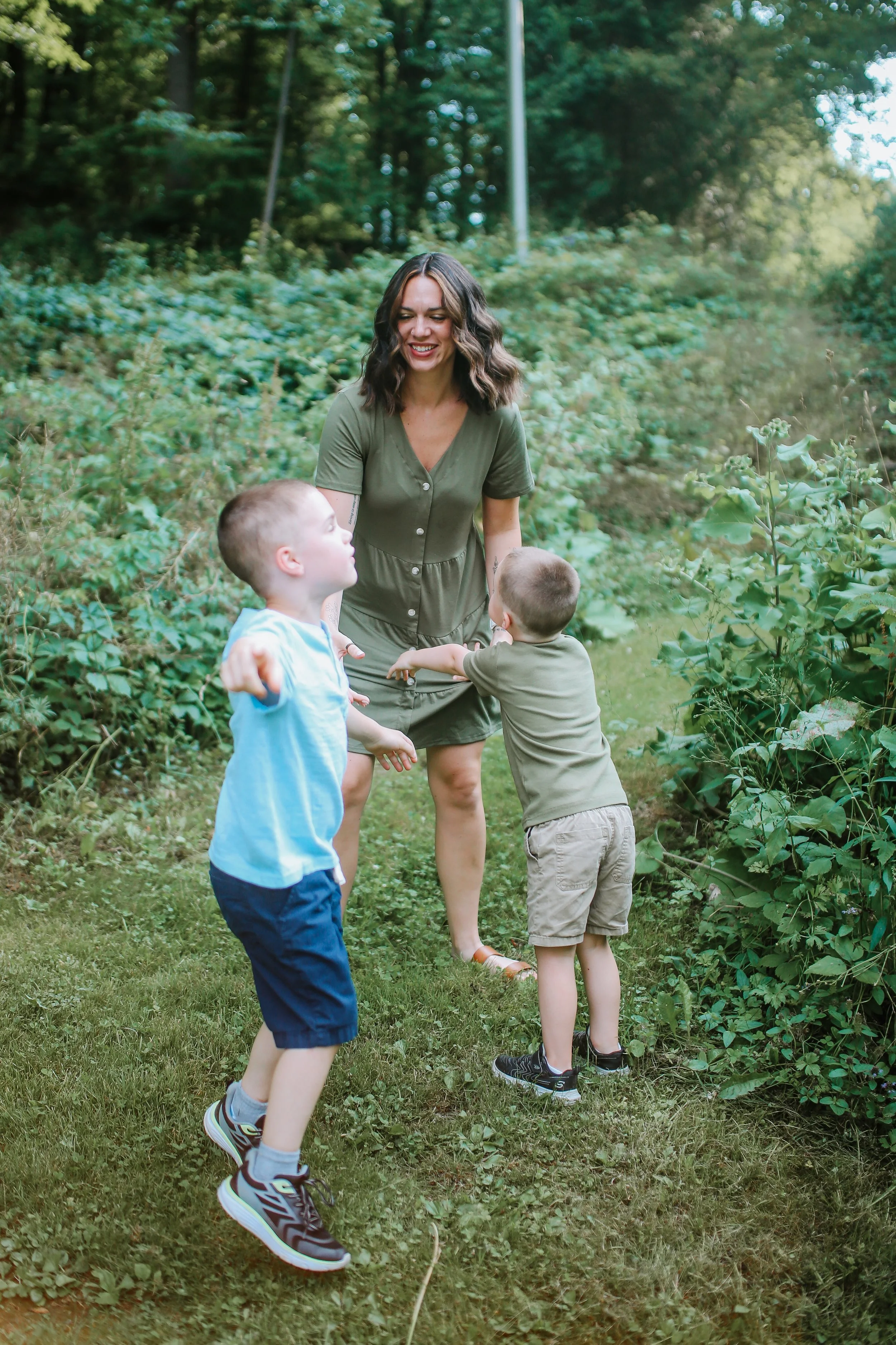 A woman playing with two young boys in a wooded outdoor area, smiling and laughing.